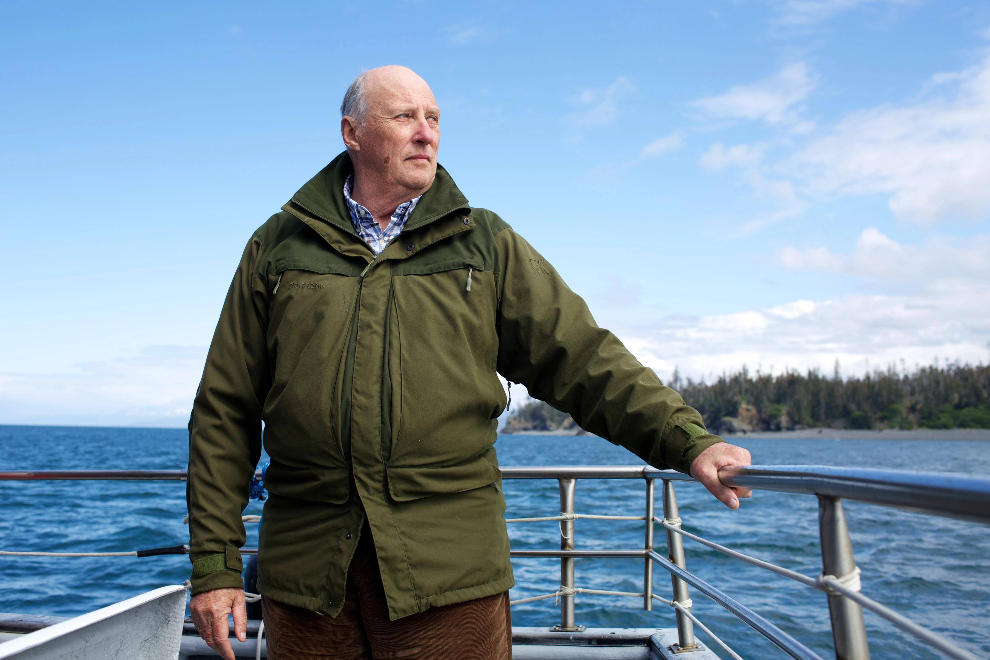 Kong Harald i kledd grønn ytterjakke, stående mens han holder seg i et rekkverk.