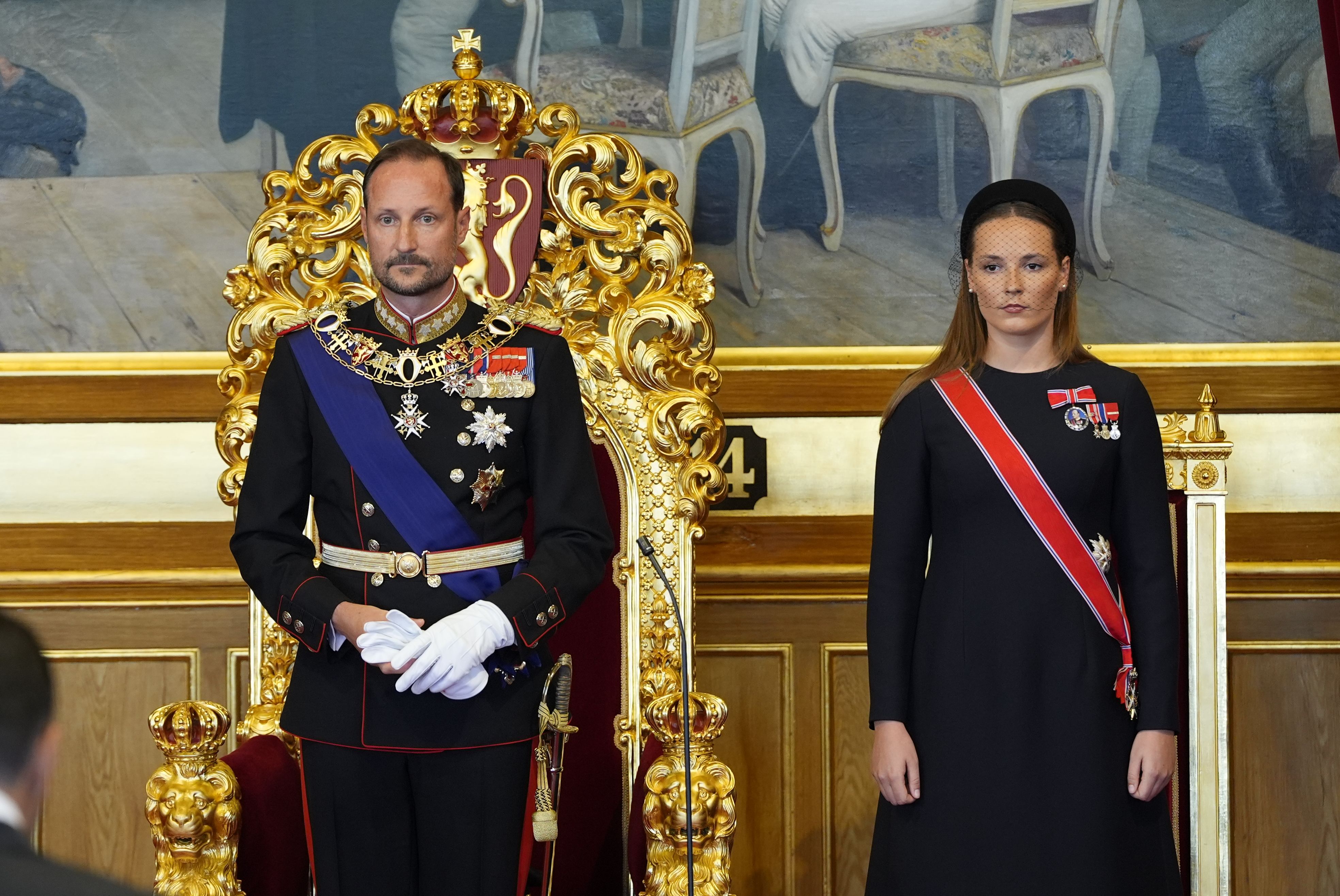 Kongen står foran tronstolen. Kronprinsessen står ved hans venstre side. Han er kledd i gallauniform, hun i sort kjole med ordensbånd.