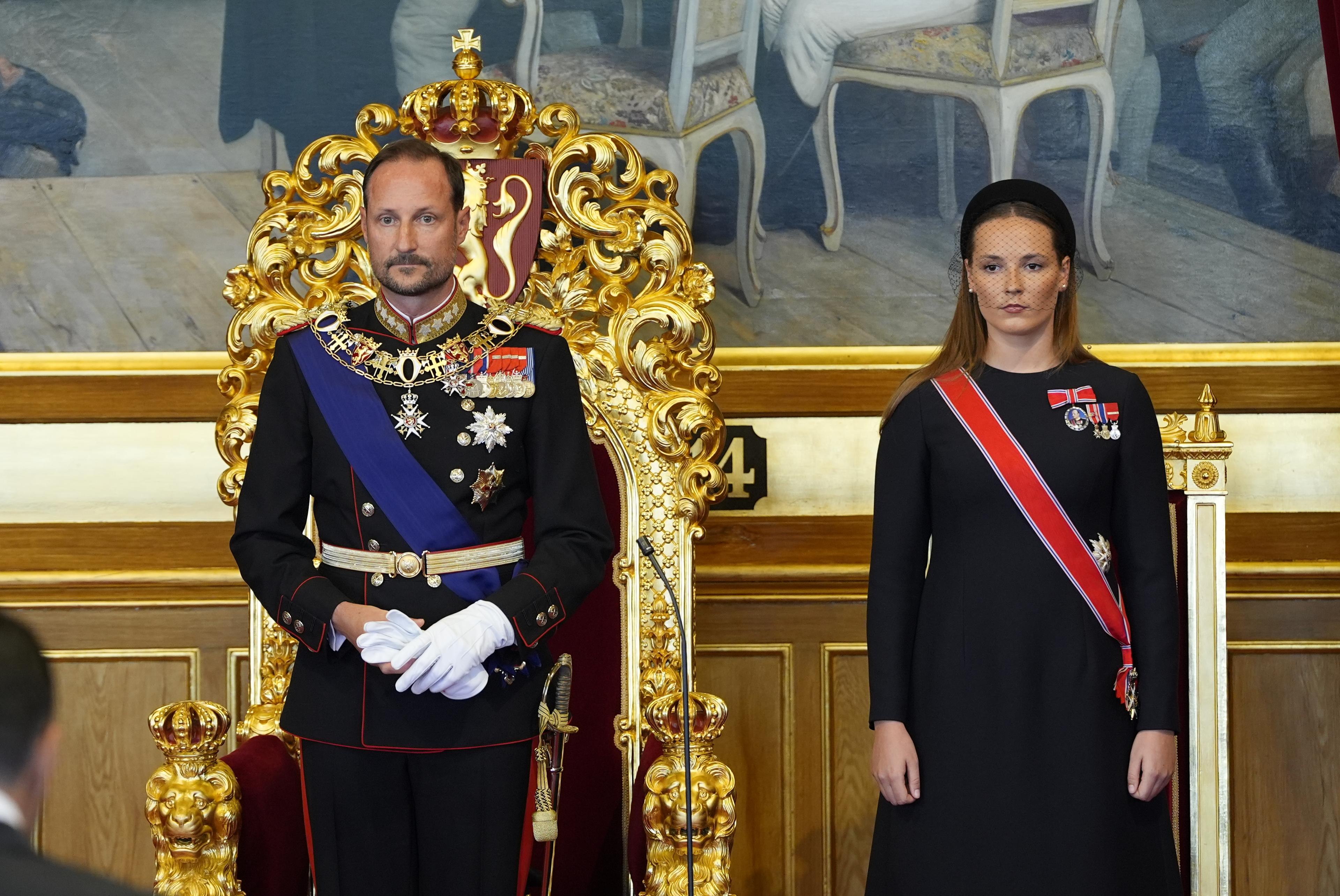 Kongen står foran tronstolen. Kronprinsessen står ved hans venstre side. Han er kledd i gallauniform, hun i sort kjole med ordensbånd.