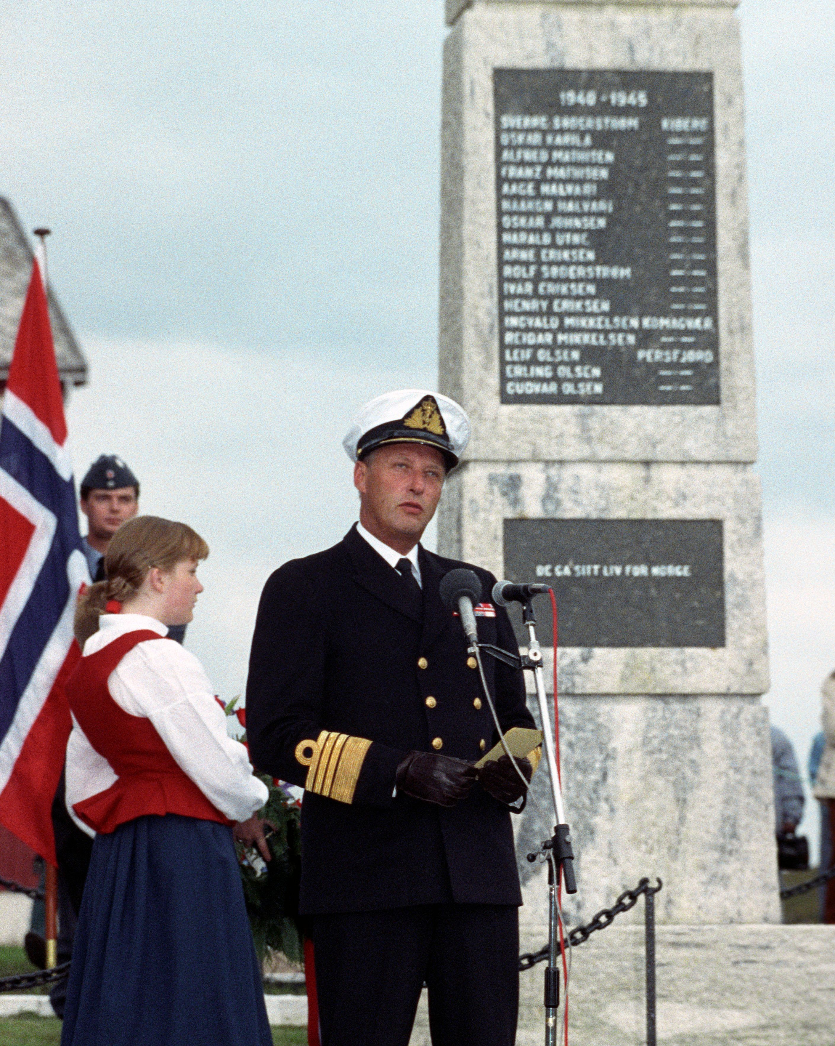 Kongen står i marineuniform ved siden av bautaen med de falnes navn.