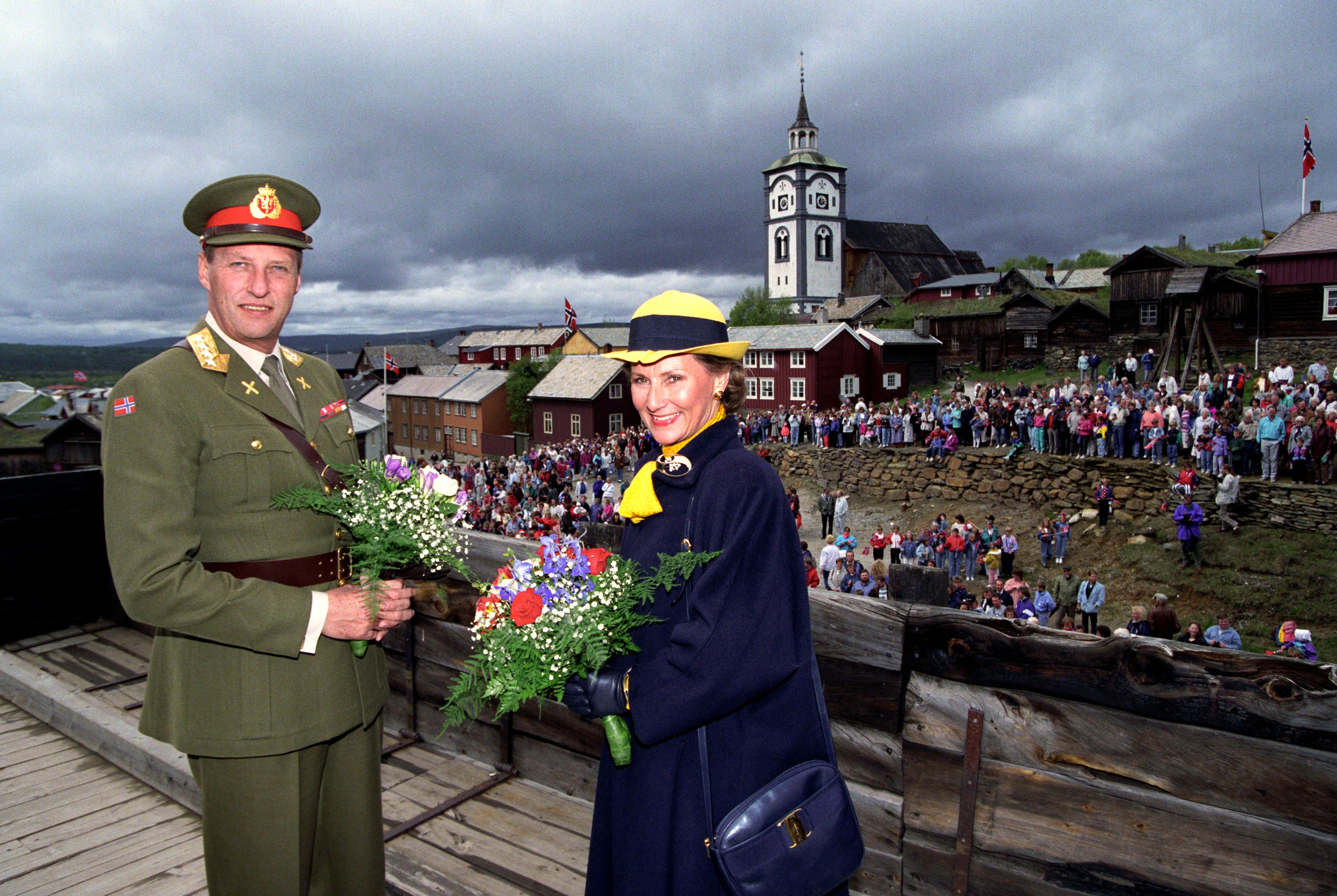 Kongeparet med blomster hendene. Bak ser vi mange frammøtte og det karakteristiske tårnet på Røros kirke.