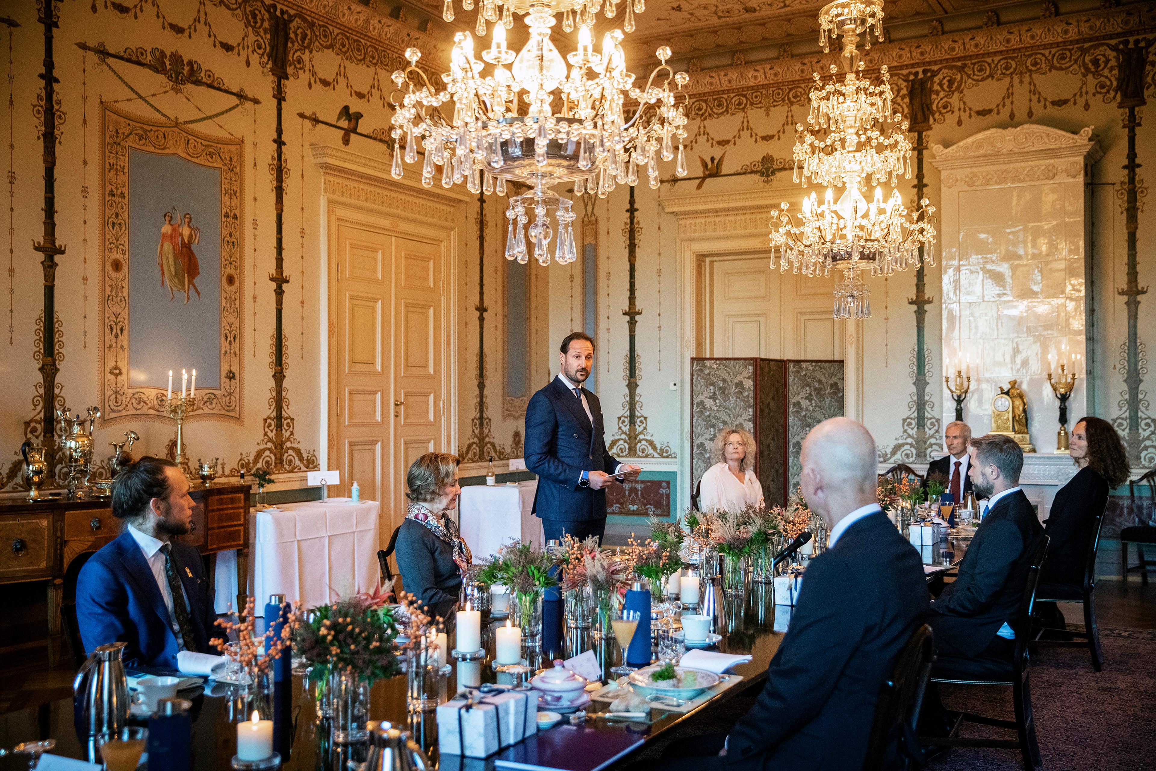 Kronprinsen står ved bordet og holder tale. Helseminister Bent Høie, Camilla Stoltenberg fra Folkehelseinsituttet med flere lytter til Kronprinsen. Sitter rundt et dekket bord i Daglige spisesal.