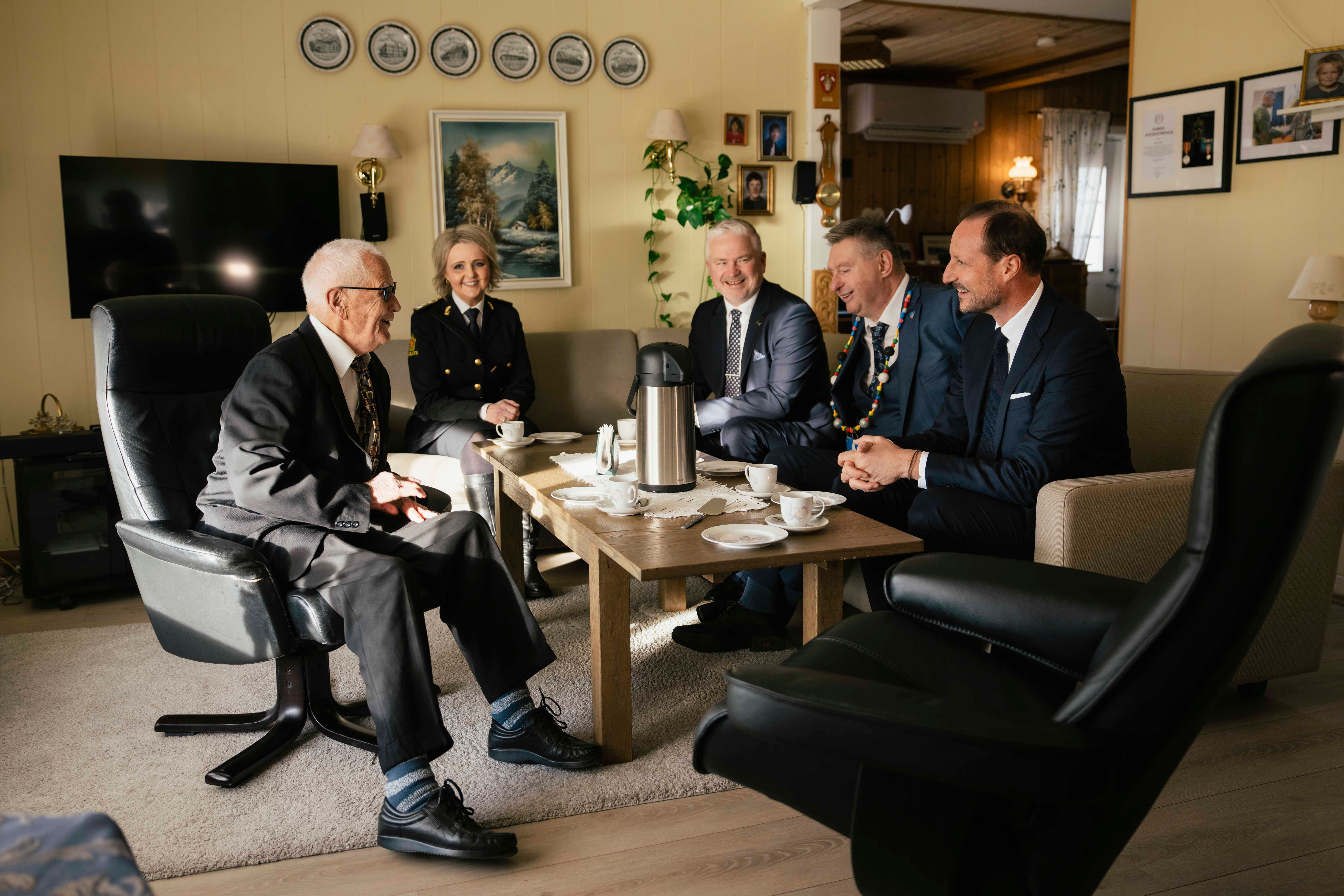 Kongen og Øymo sitter rundt kaffebordet sammen med andre gjester i stua hjemme hos Øymo.