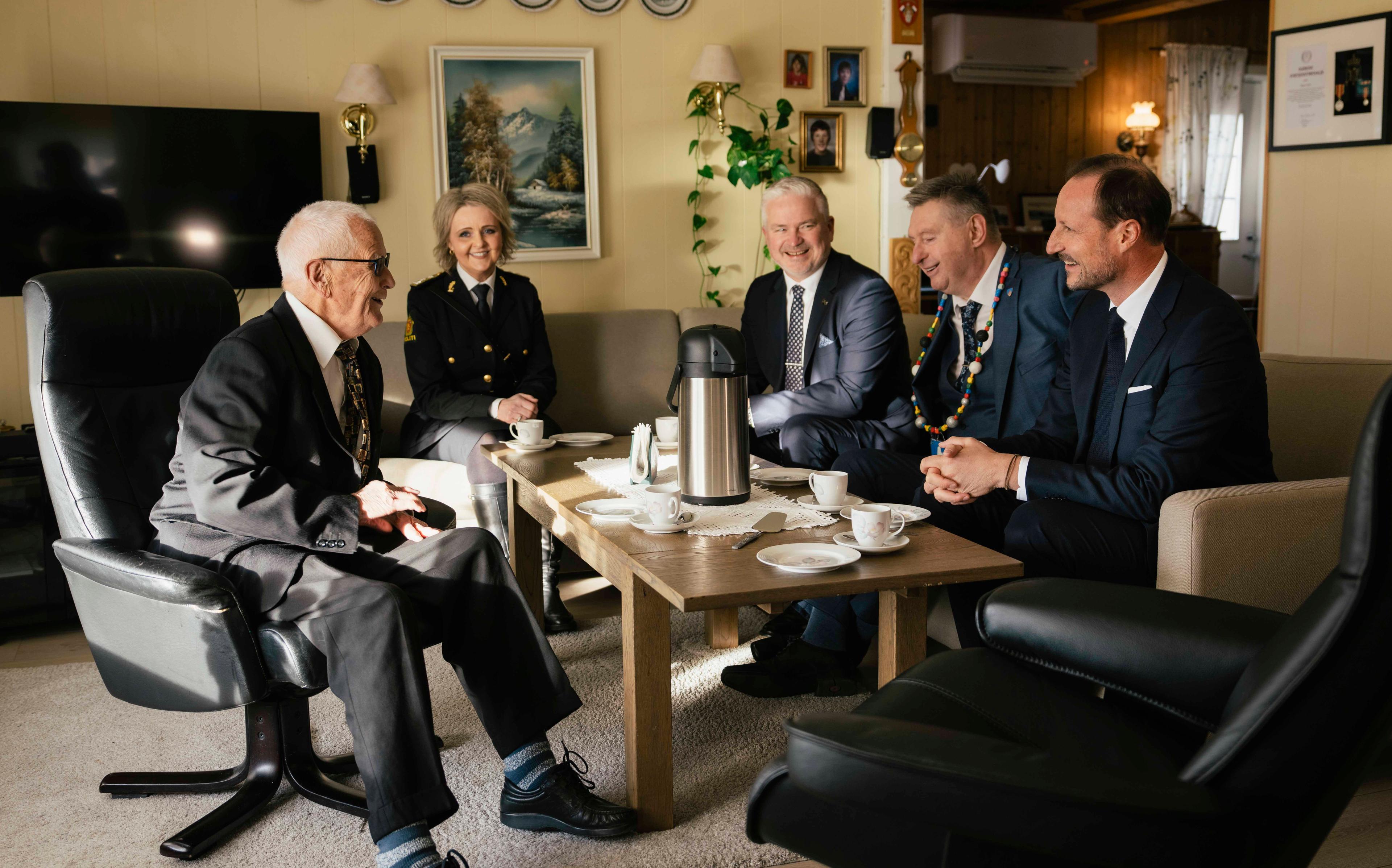 Kongen og Øymo sitter rundt kaffebordet sammen med andre gjester i stua hjemme hos Øymo.