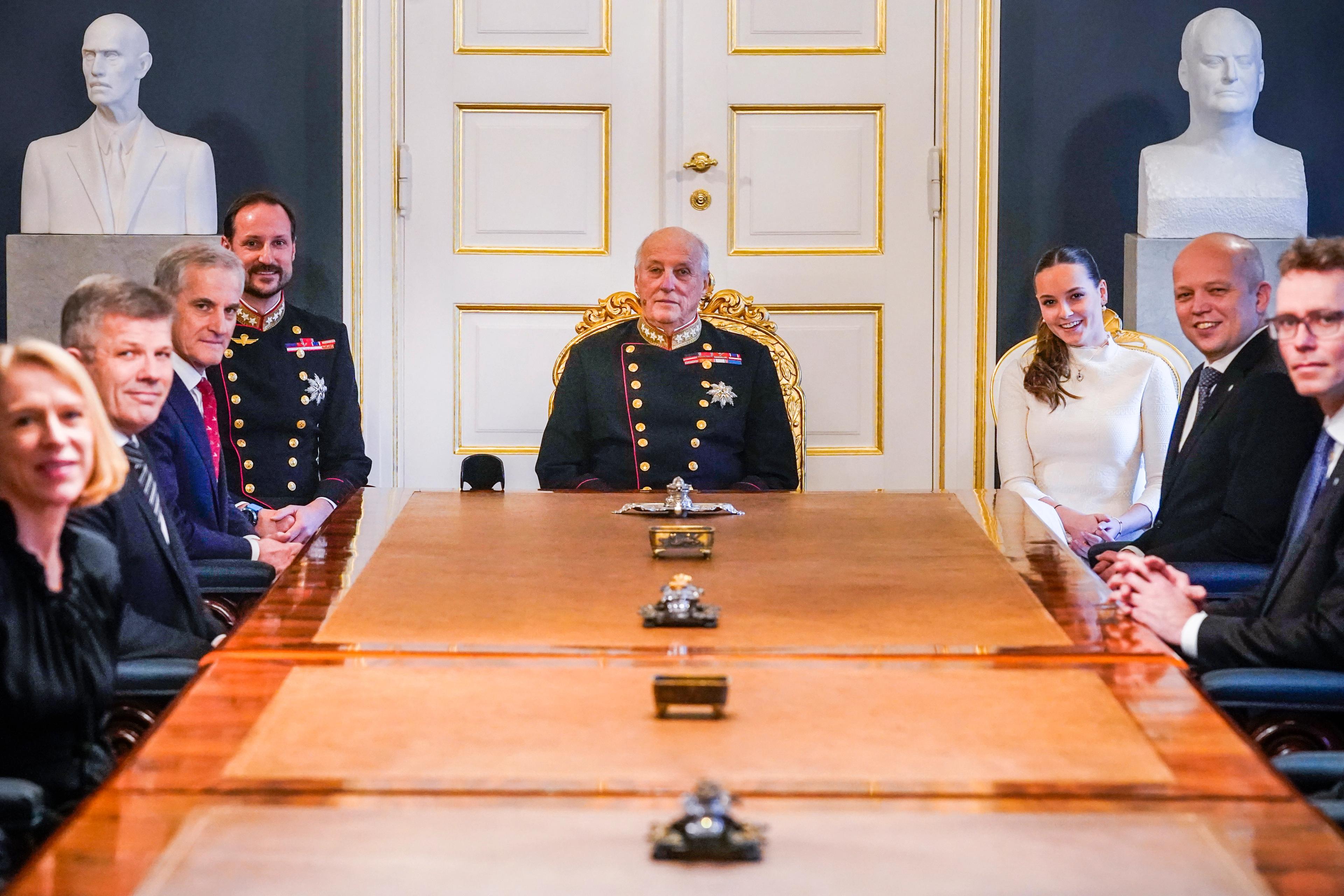 Kong Harald sitter i gallauniform med Kronprins Haakon og Prinsesse Ingrid Alexandra på hver side. Bak ham står bystene av Kong Haakon VII og Kong Olav. Ulike regjeringsmedlemmer fyller stolene nedover langs bordet.
