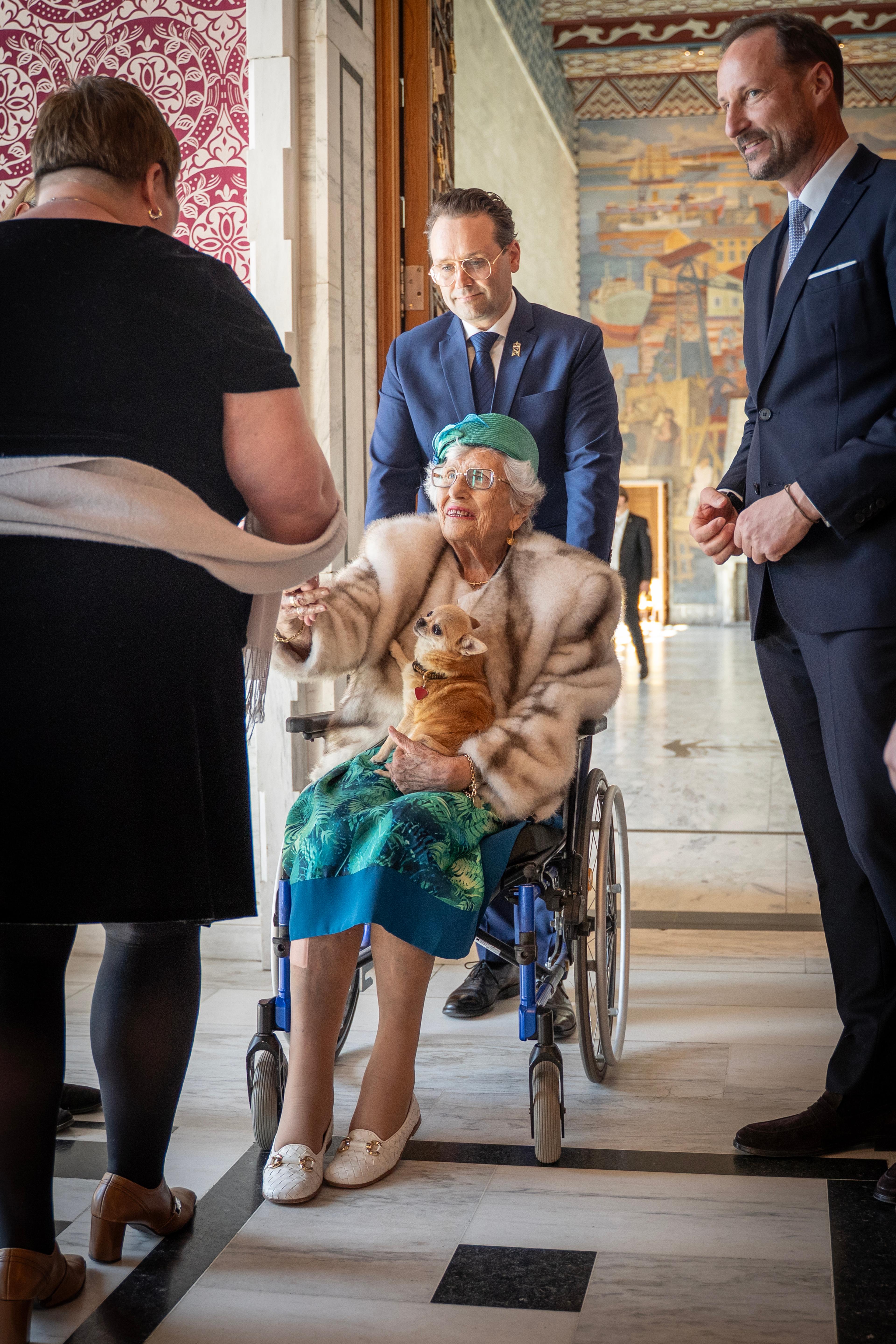 Prinsesse Astrid sitter i en rullestol med Wilma på fanget og rekker fram en hånd mot Kathrin Dahlstrøm, som står med ryggen til.