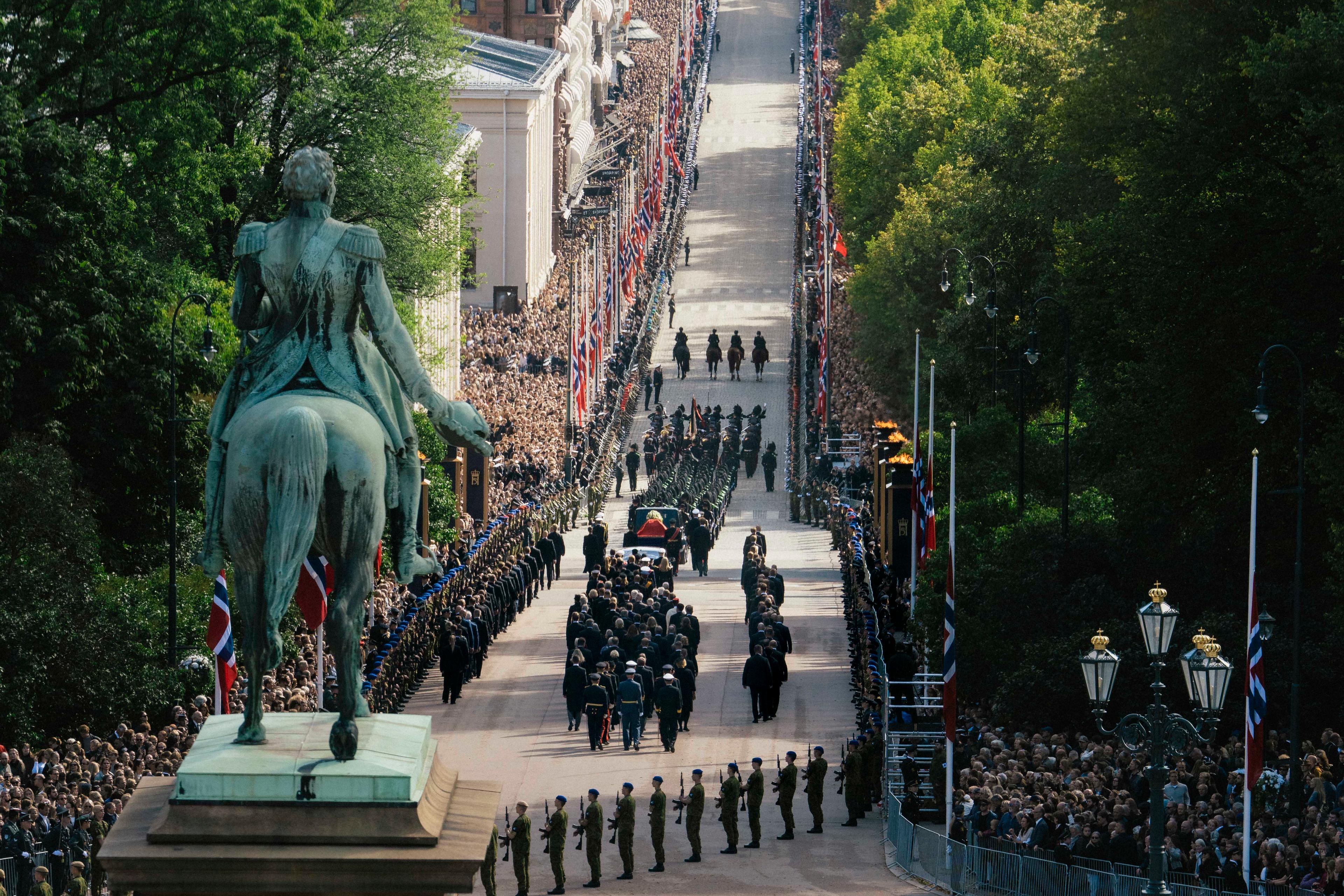 Prosesjonen på vei ned Slottsbakken, omkranset av mange mennesker og flagg på halv stang. Soldater står i giv akt langs ruten. I forgrunnen ligger et hav av blomster rundt statuen av Kong Carl Johan.