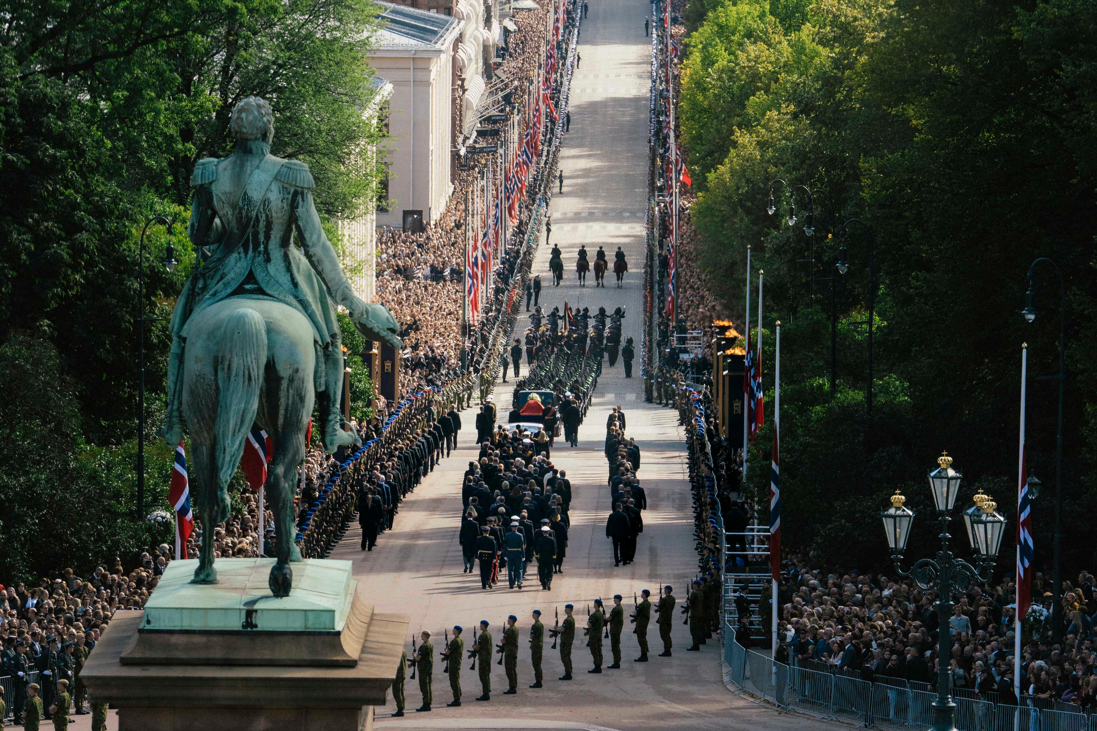 Prosesjonen på vei ned Slottsbakken, omkranset av mange mennesker og flagg på halv stang. Soldater står i giv akt langs ruten. I forgrunnen ligger et hav av blomster rundt statuen av Kong Carl Johan.