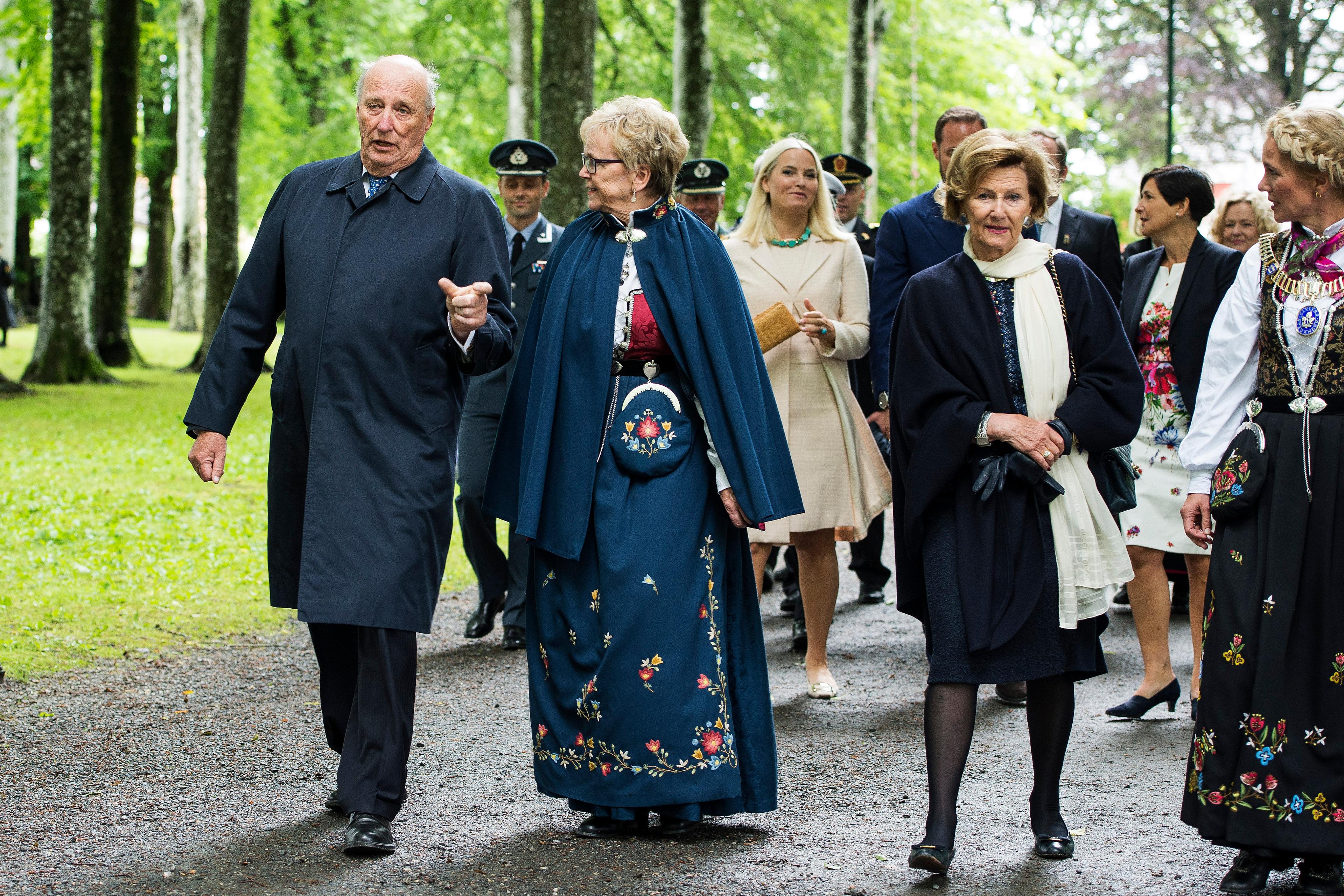 Kong Harald og Dronning Sonja kommer gående nedover en grusvei, ledsaget av statsforvalteren i Rogaland. Kronprinsparet og en rekke andre følger på.