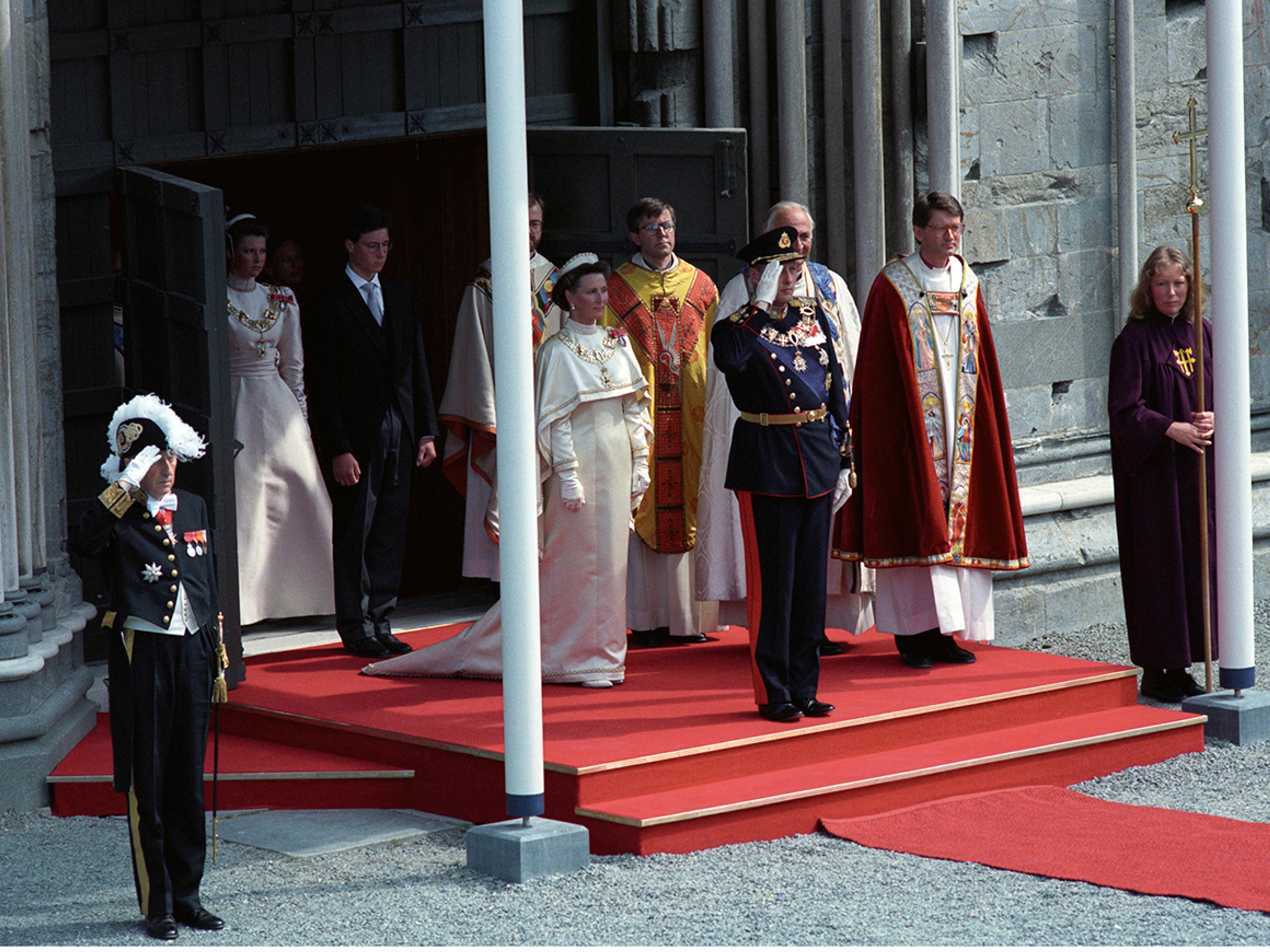 Kongen og Dronningen står på trappen, som er dekket med rød løper. Kongen gjør honnør.