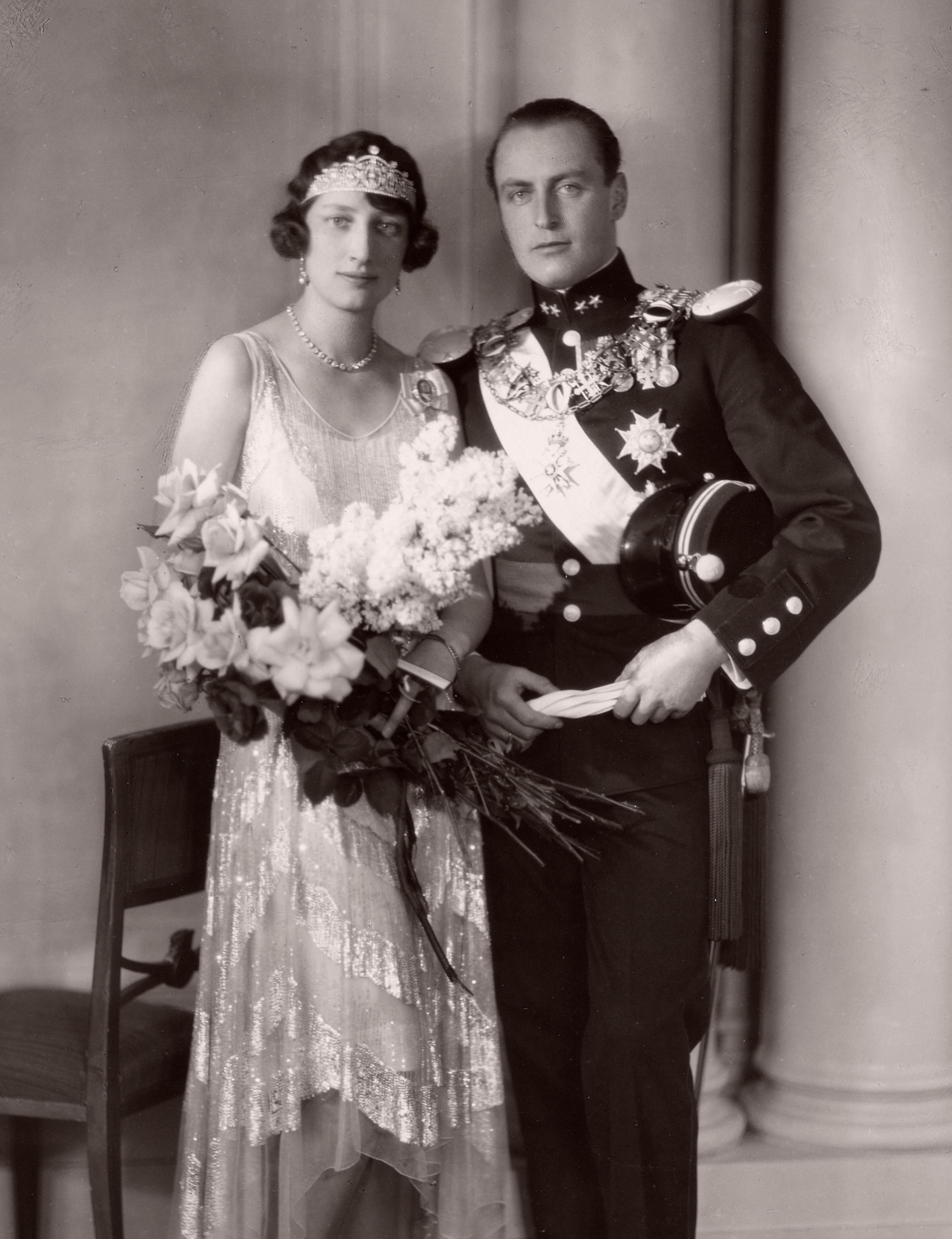 Kronprins Olav i gallauniform og Prinsesse Märtha med diadem og lys kjole. Hun har armene fulle av blomster.
