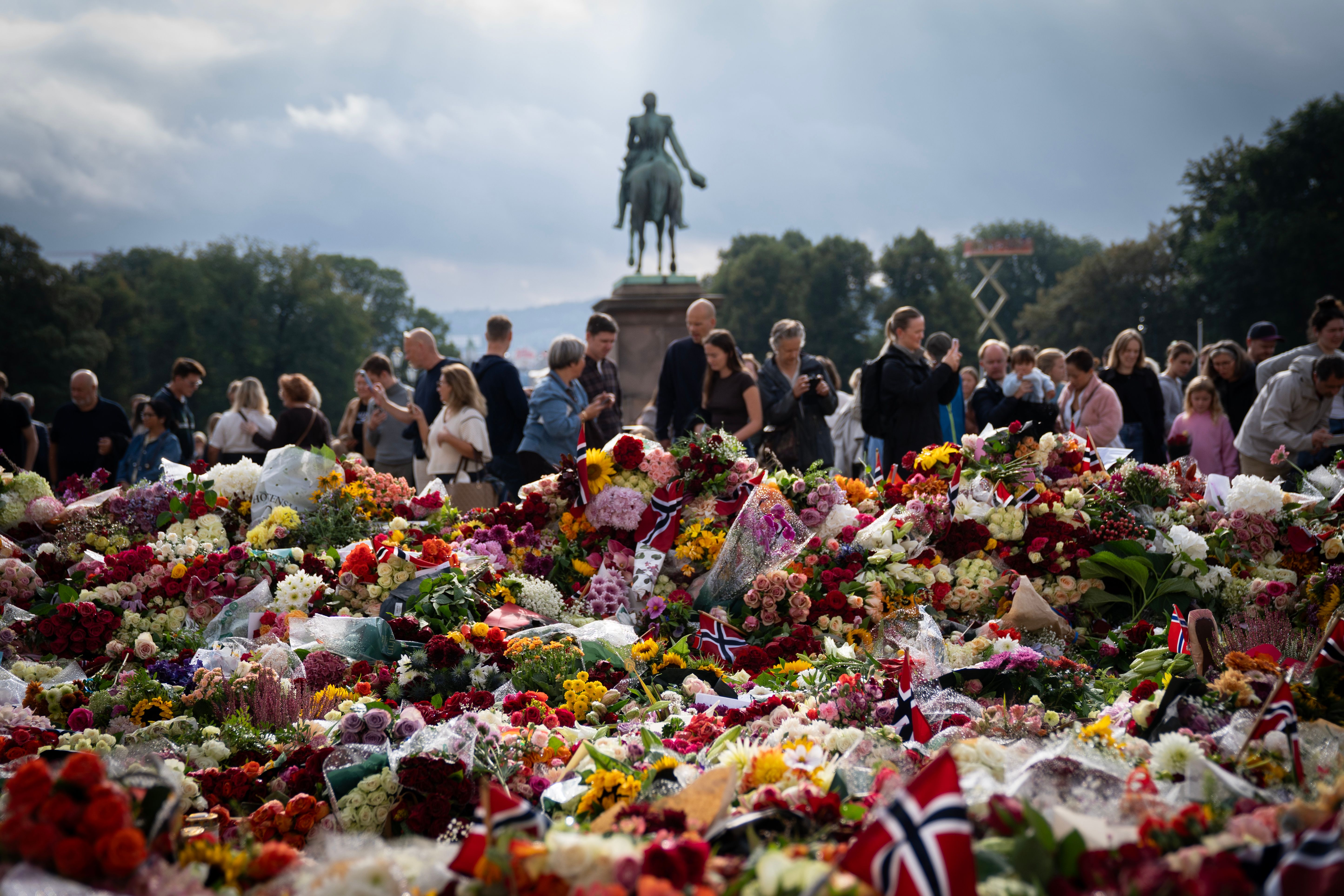 Forgrunnen er fylt av blomster og flagg. I bakgrunnen ser man Karl Johan-statuen og en rekke mennesker som står rundt blomsterhavet.