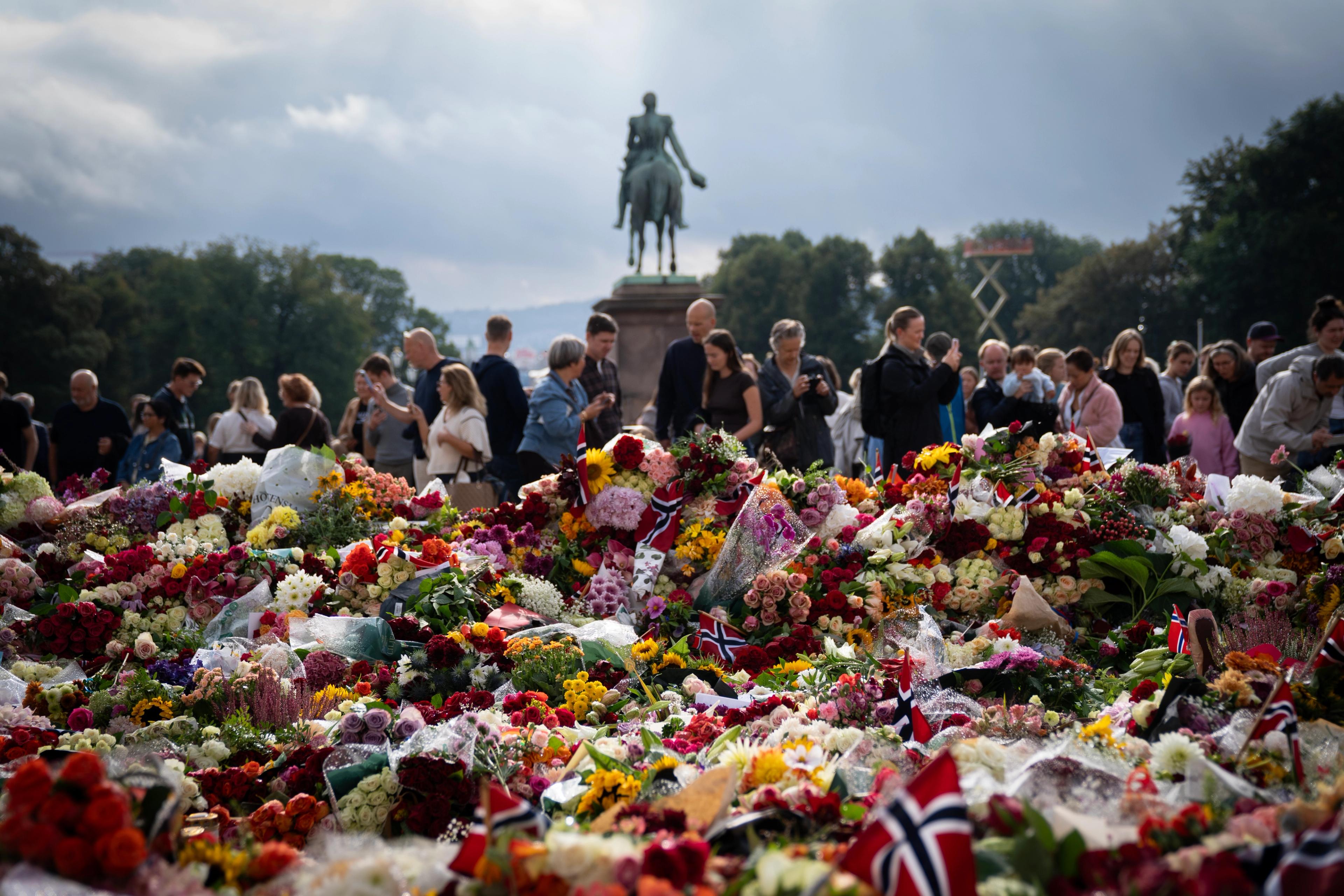 Forgrunnen er fylt av blomster og flagg. I bakgrunnen ser man Karl Johan-statuen og en rekke mennesker som står rundt blomsterhavet.