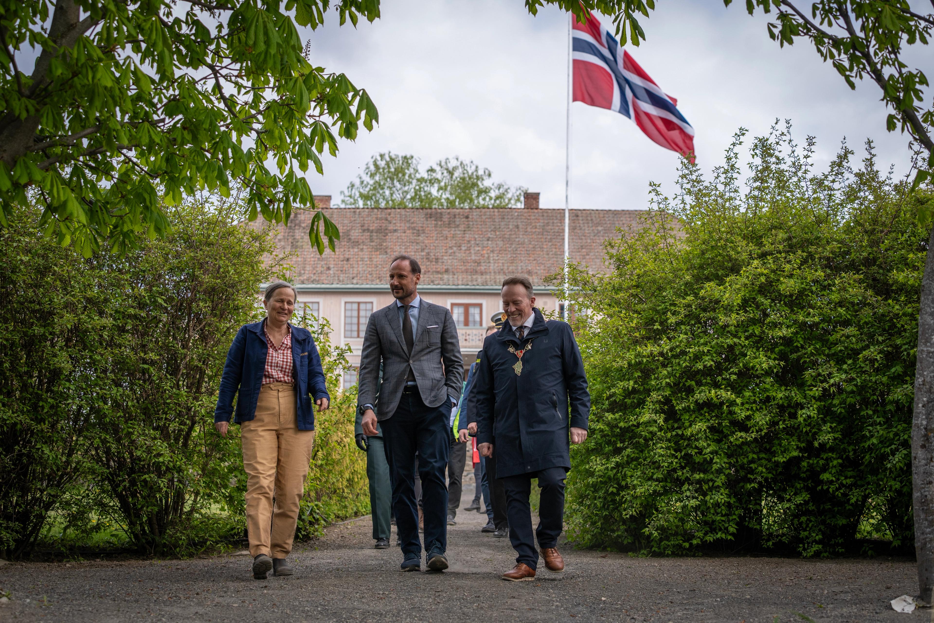 De tre går sammen ned en allé, med det norske flagget heist ved hovedhuset i bakgrunnen.