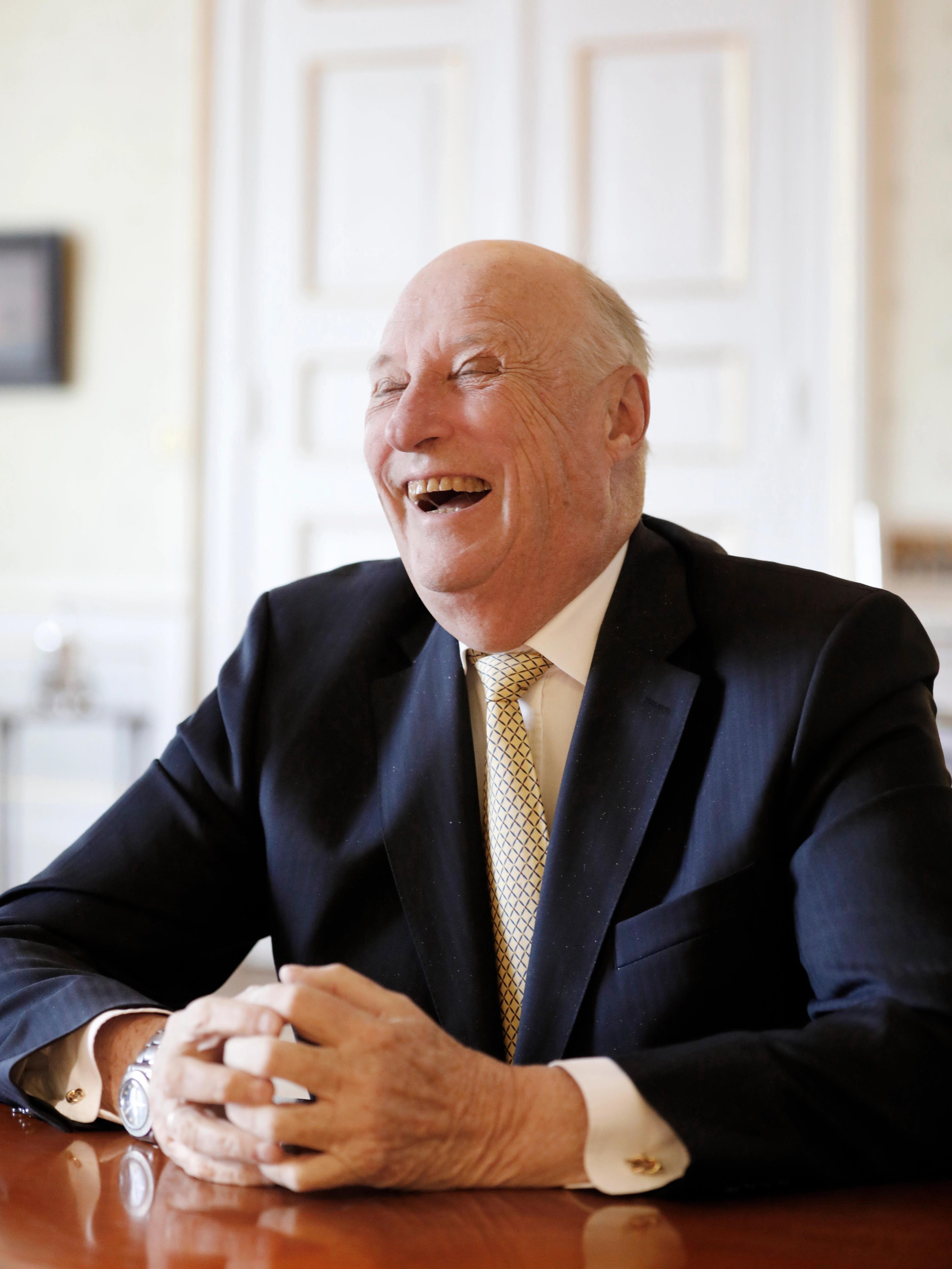 Kong Harald ler mens han sitter med hendene foldet på bordet foran seg.