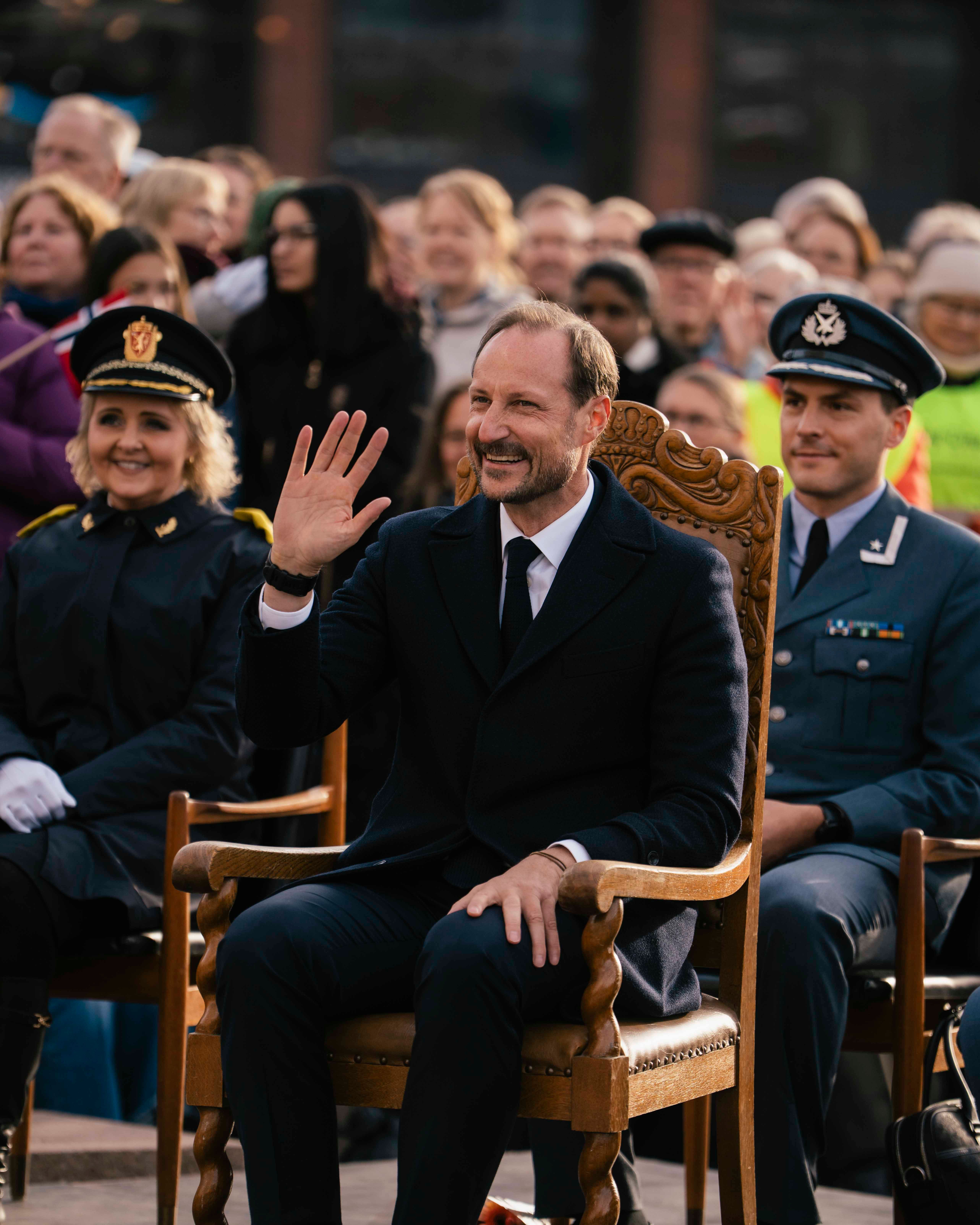Kongen sitter på en stol og vinker. Bak han ser vi en folkemenge