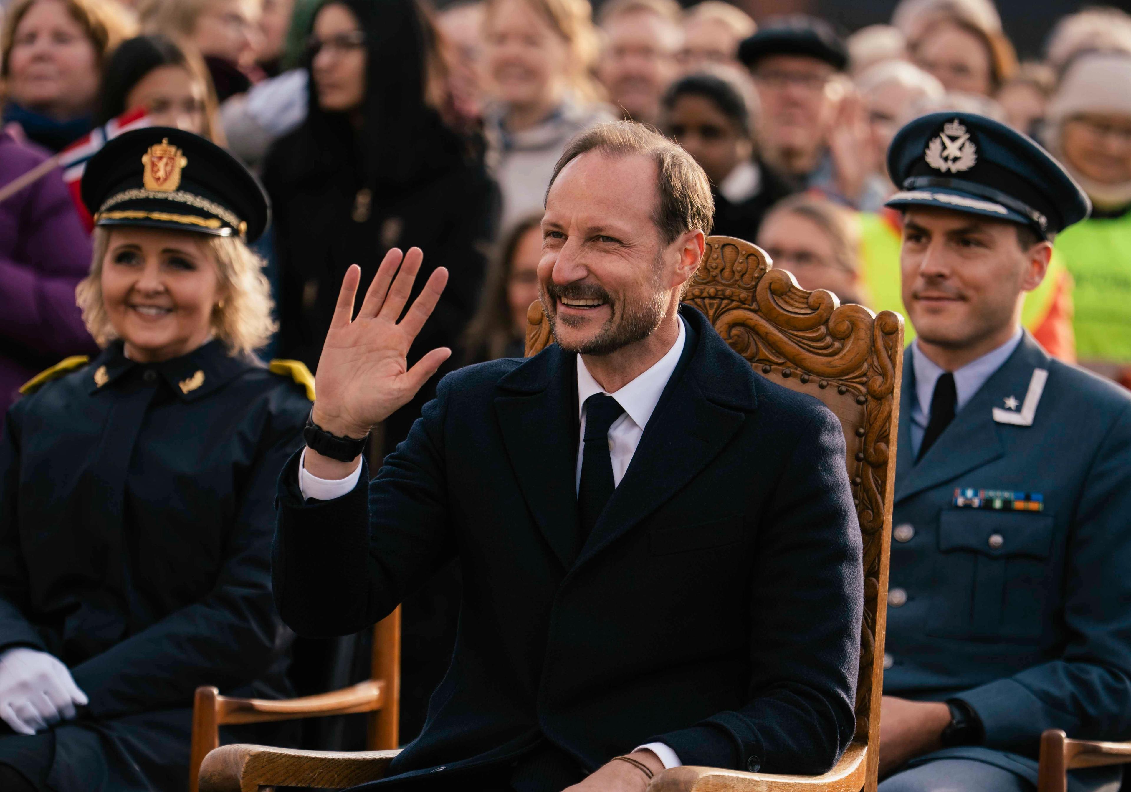Kongen sitter på en stol og vinker. Bak han ser vi en folkemenge