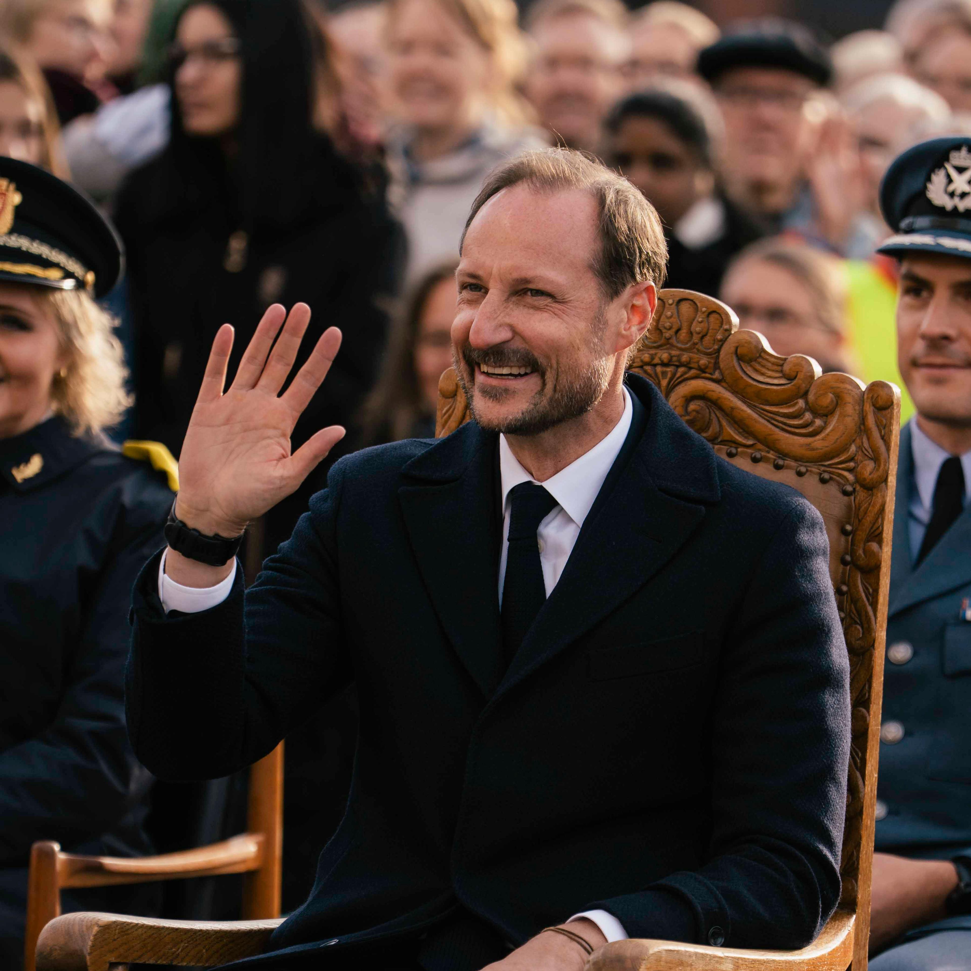 Kongen sitter på en stol og vinker. Bak han ser vi en folkemenge