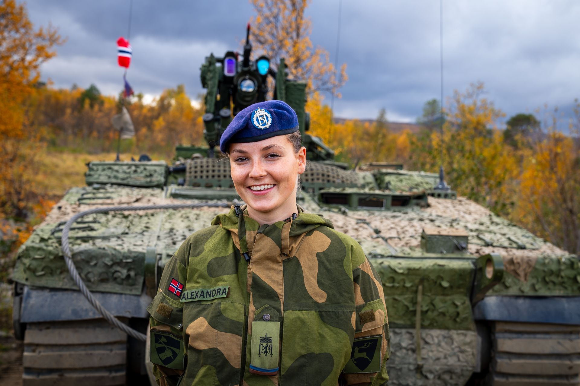 Prinsessen står i feltuniform med blå beret foran en kamuflasjefarget tanks.
