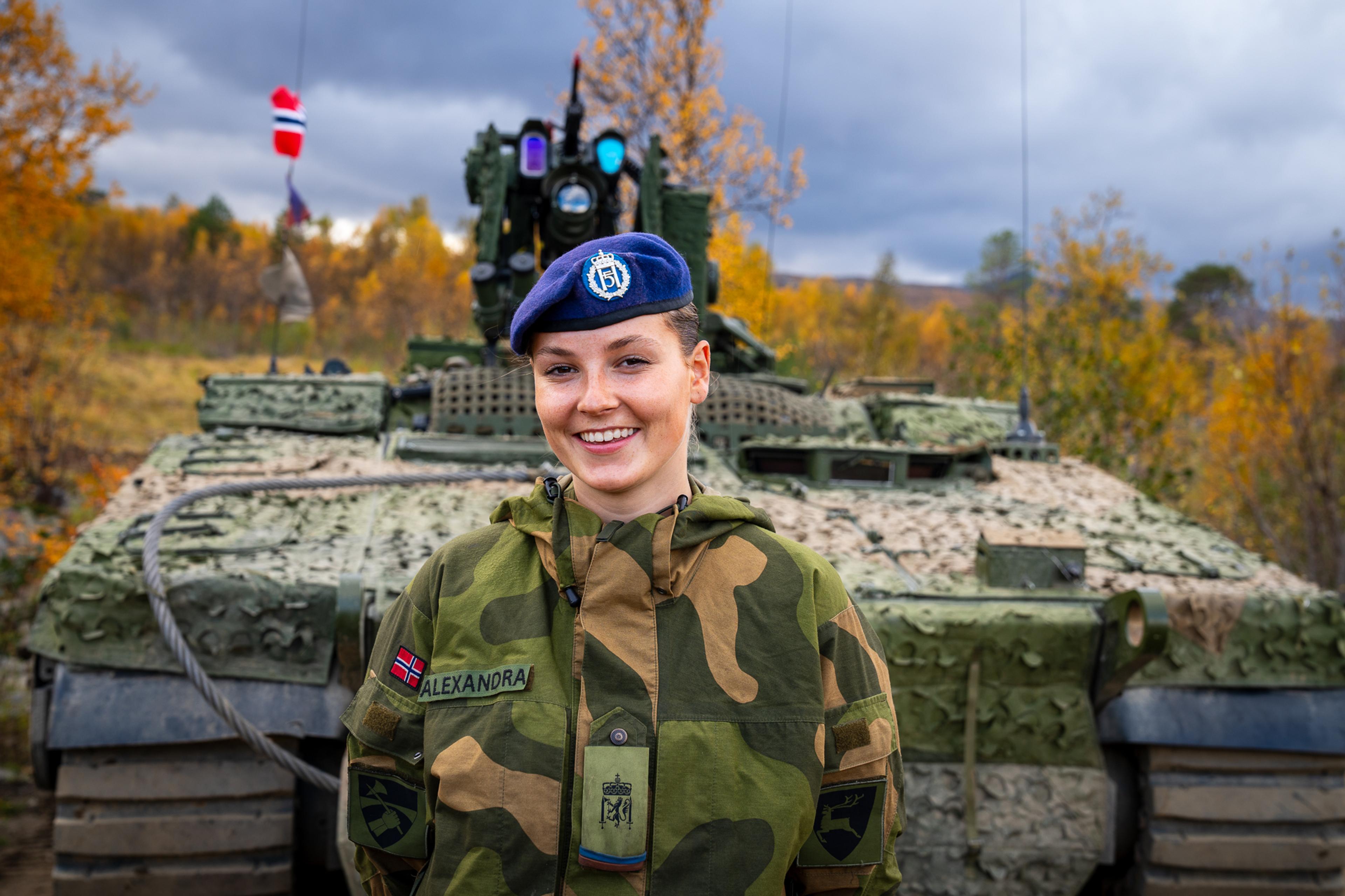Prinsessen står i feltuniform med blå beret foran en kamuflasjefarget tanks.