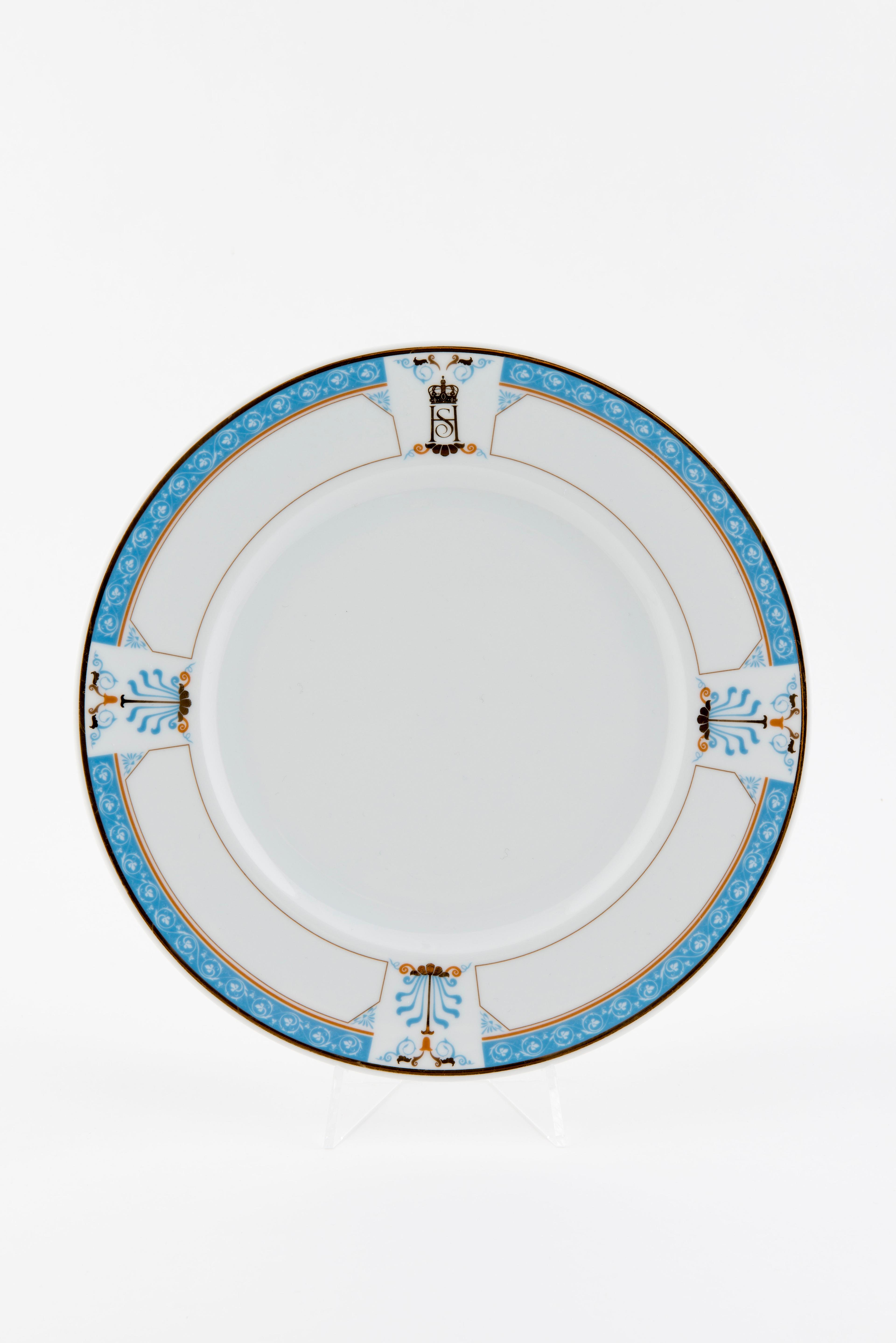 En hvit tallerken med en smal kant med blå dekor. Kanten har også tre felt med større palmetter i tillegg til Kongeparets fellesmonogram.