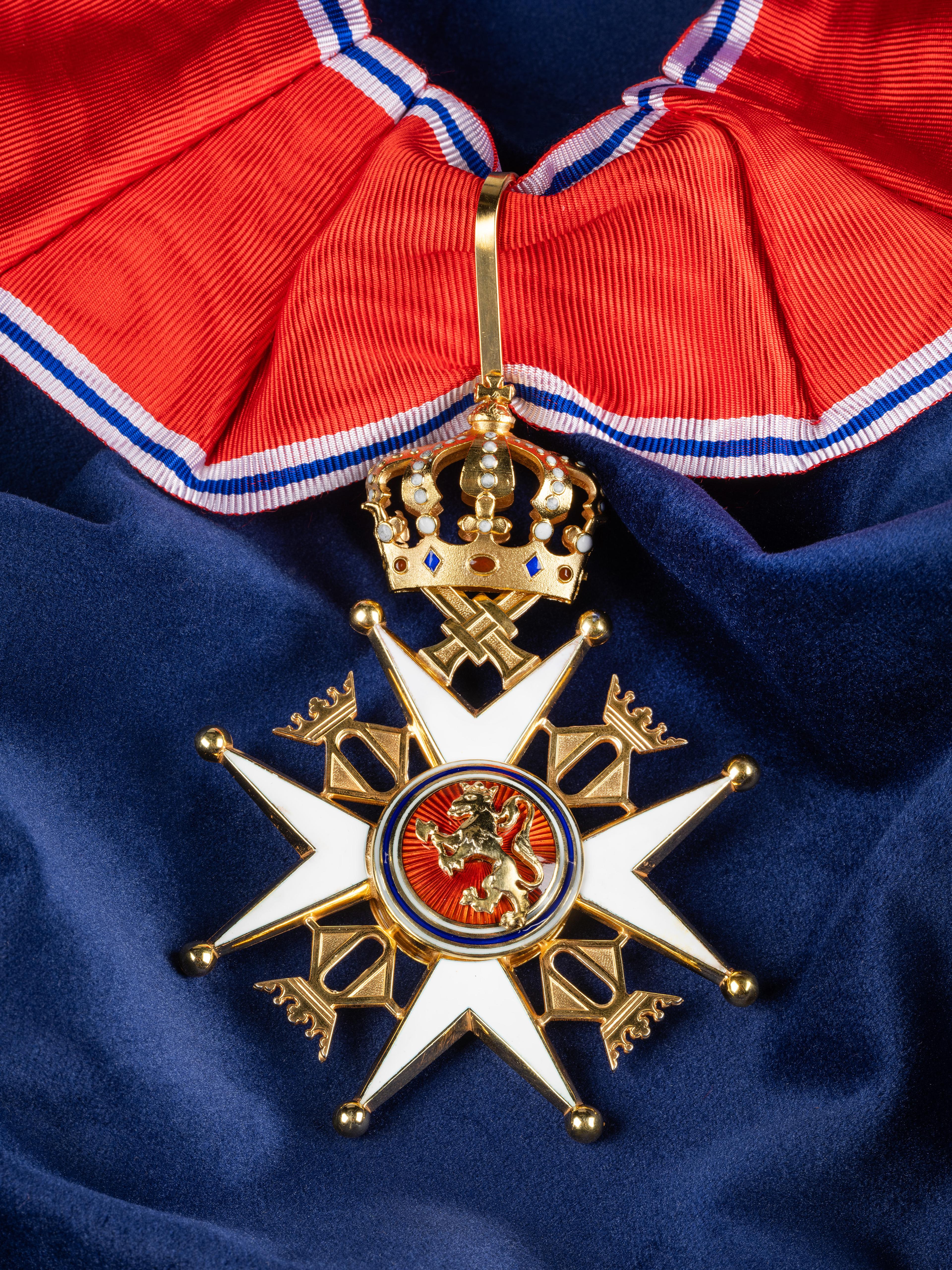 Nærbilde av ordensmerket festet i rødt bånd med blå og hvite striper på en bakgrunn av blått klede.