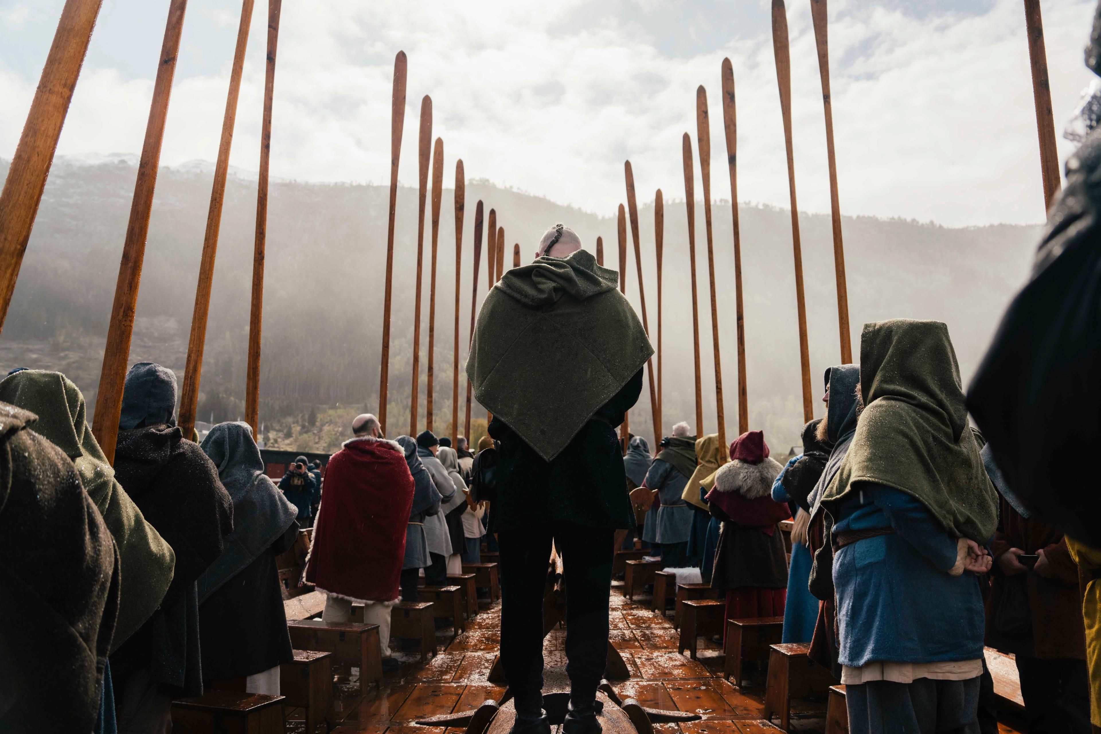 En mann kledd i vikingklær står med ryggen til midt i et stort vikingskip. På hver side av han sitter det en rad med folk som peker med åren til værs. I bakgrunnen ser man fjell og solglimt gjennom skyer.