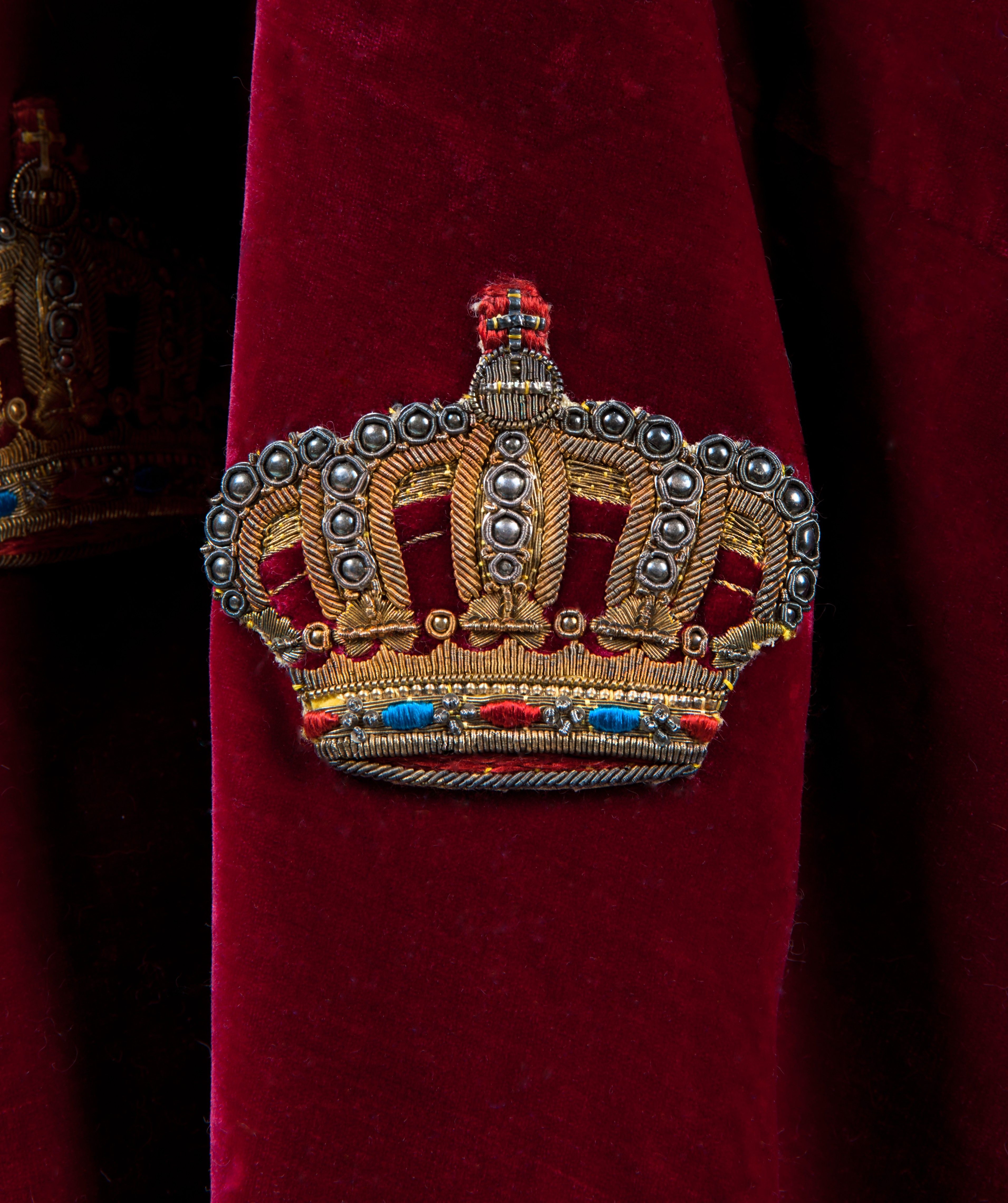 Nærbilde av en lukket krone brodert på den røde kappen med gulltråd. Blå og rød tråd illuderer juveler og det små kuler illuderer perlene på kronens buer.
