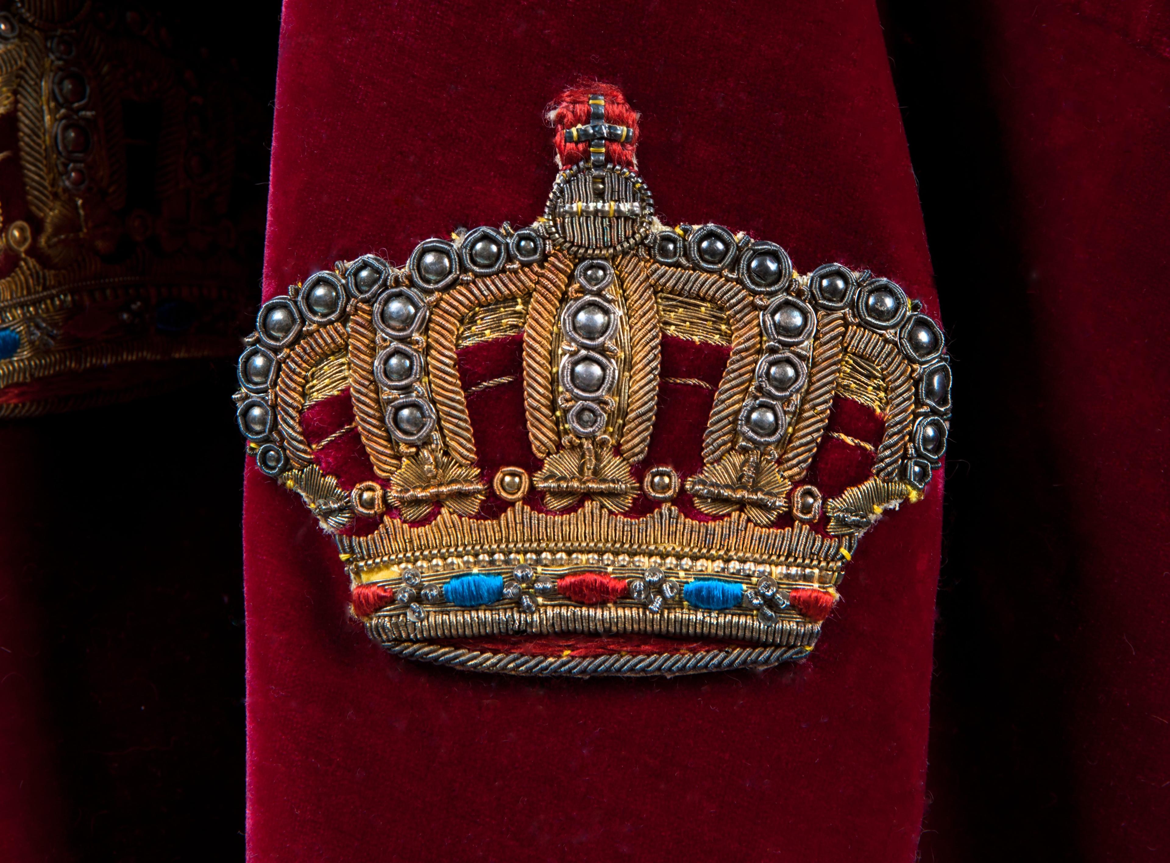 Nærbilde av en lukket krone brodert på den røde kappen med gulltråd. Blå og rød tråd illuderer juveler og det små kuler illuderer perlene på kronens buer.
