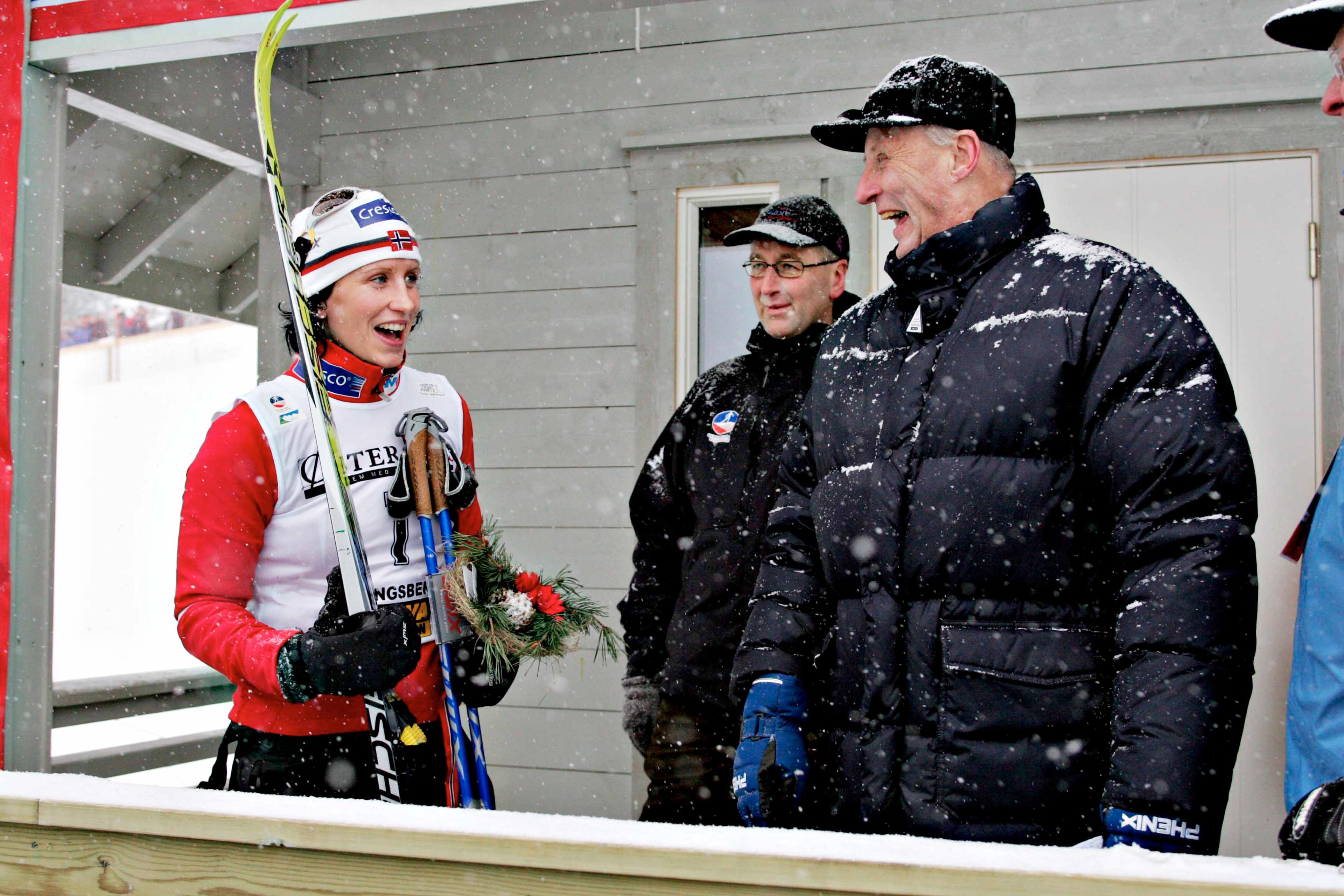 Kong Harald er ikledd sort boblejakke og ler sammen med Marit Bjørgen som står med ski, staver og langrennsantrekk. Det snør tett.
