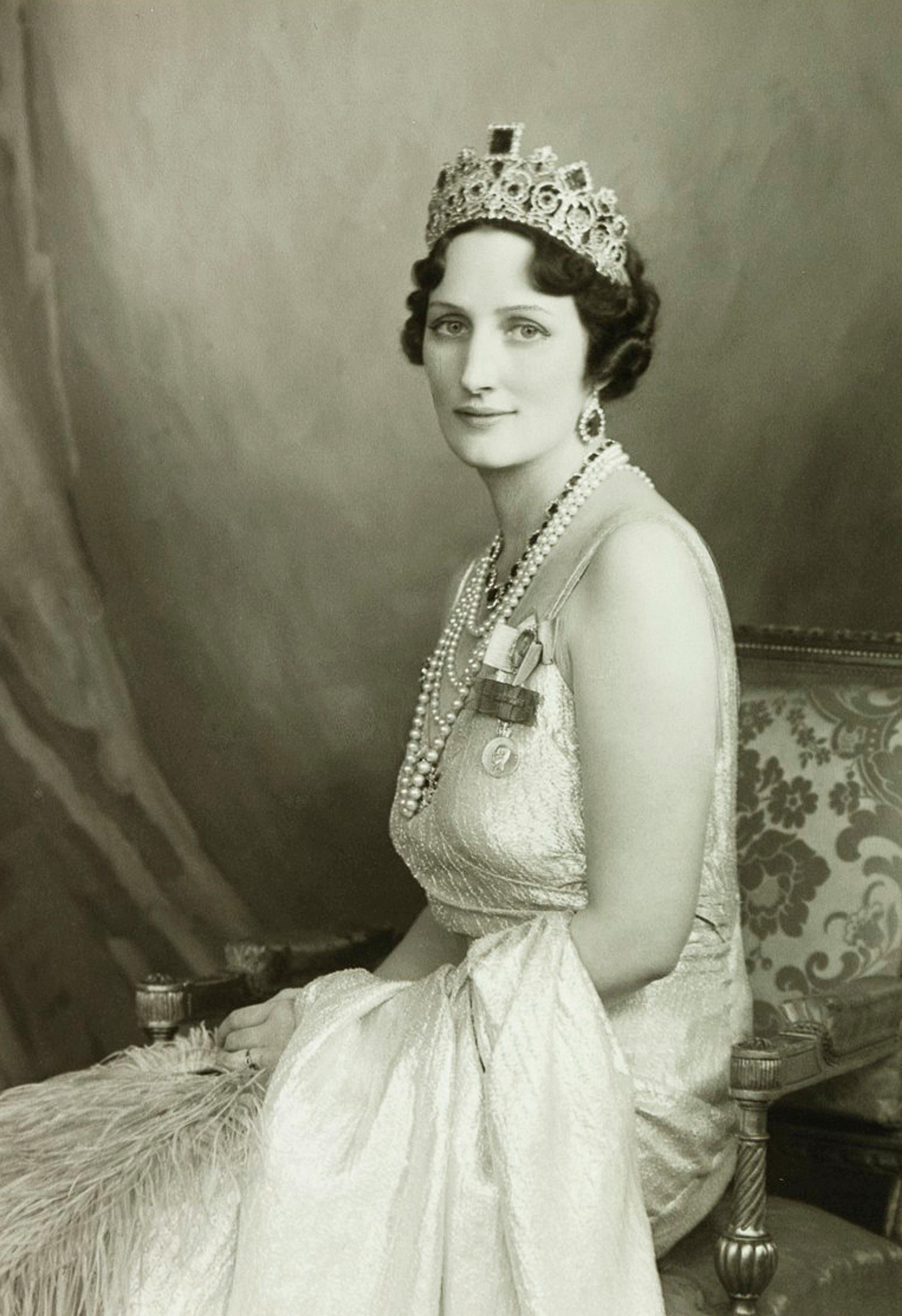 Portrett av Kronprinsesse Märtha i lys, ermeløs kjole, perlekjeder og tiara. Hunsitter på et stol med skjørtet drapert omkring seg.