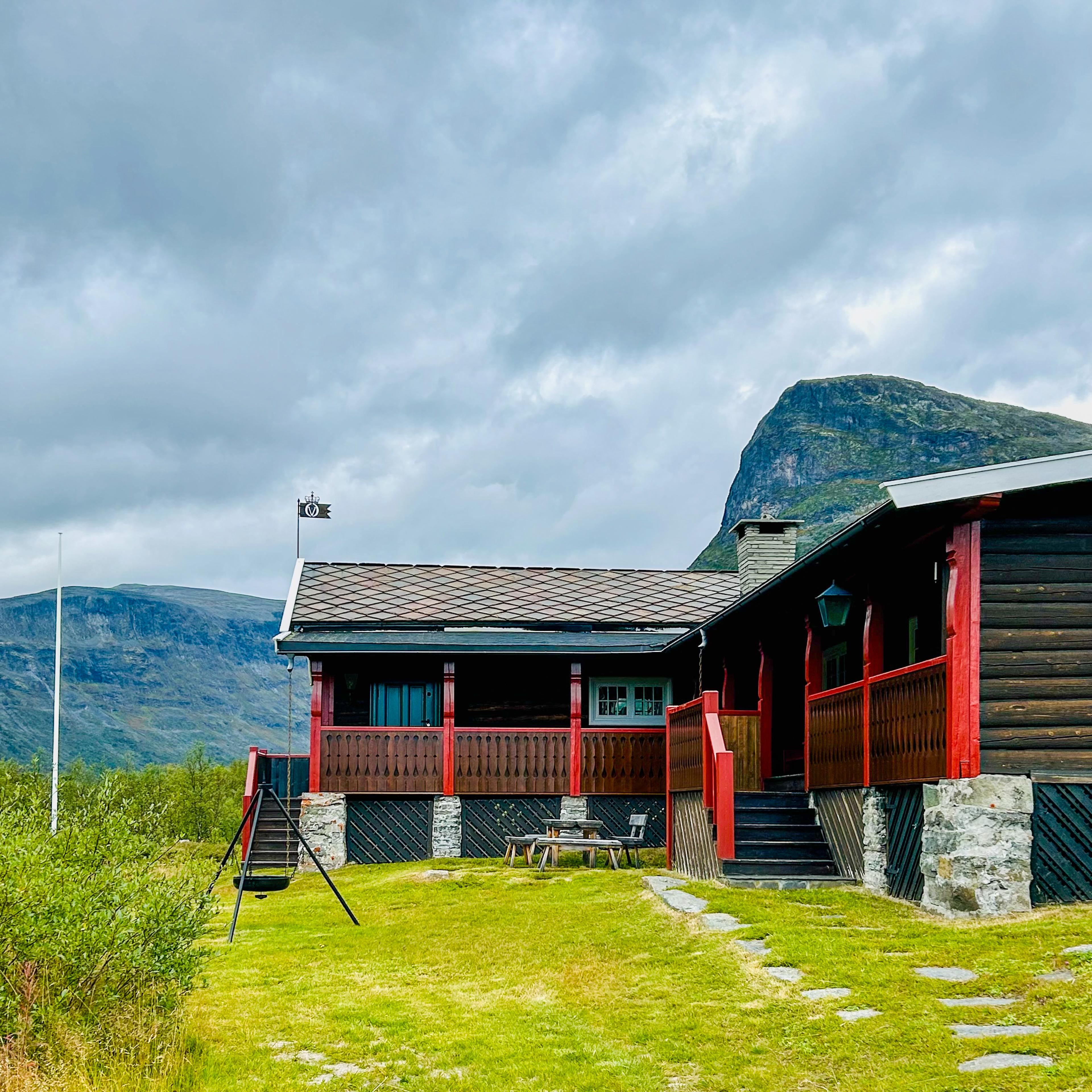 Eksteriør av en brunbeiset hytte med hvite vinduskarmer og røde stolper.