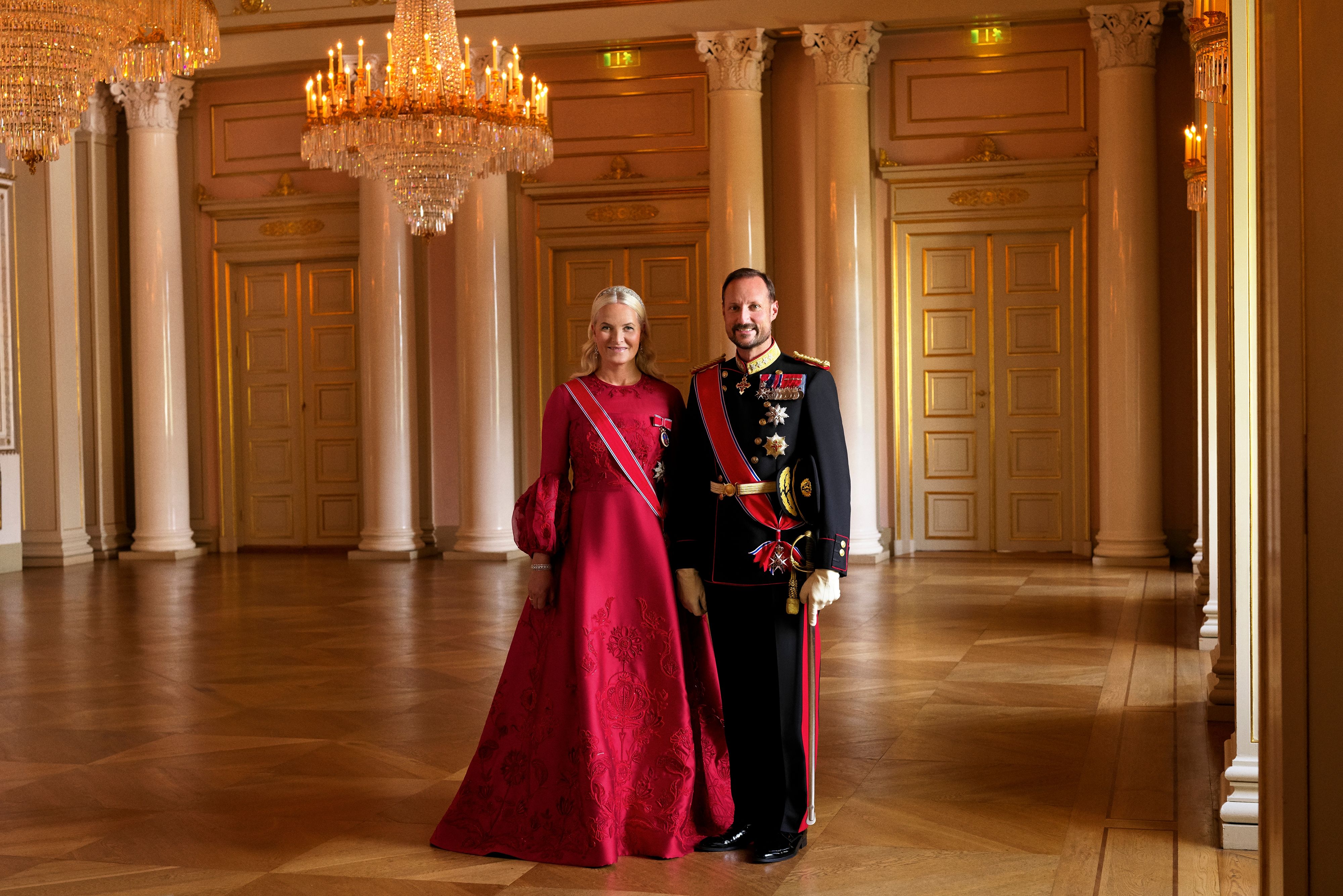 Kronprinsparet i helfigur i Store festsal. Kronprinsen har gallauniform med ordener og medaljler, Kronprinsessen har rød gallakjole og ordensbånd.