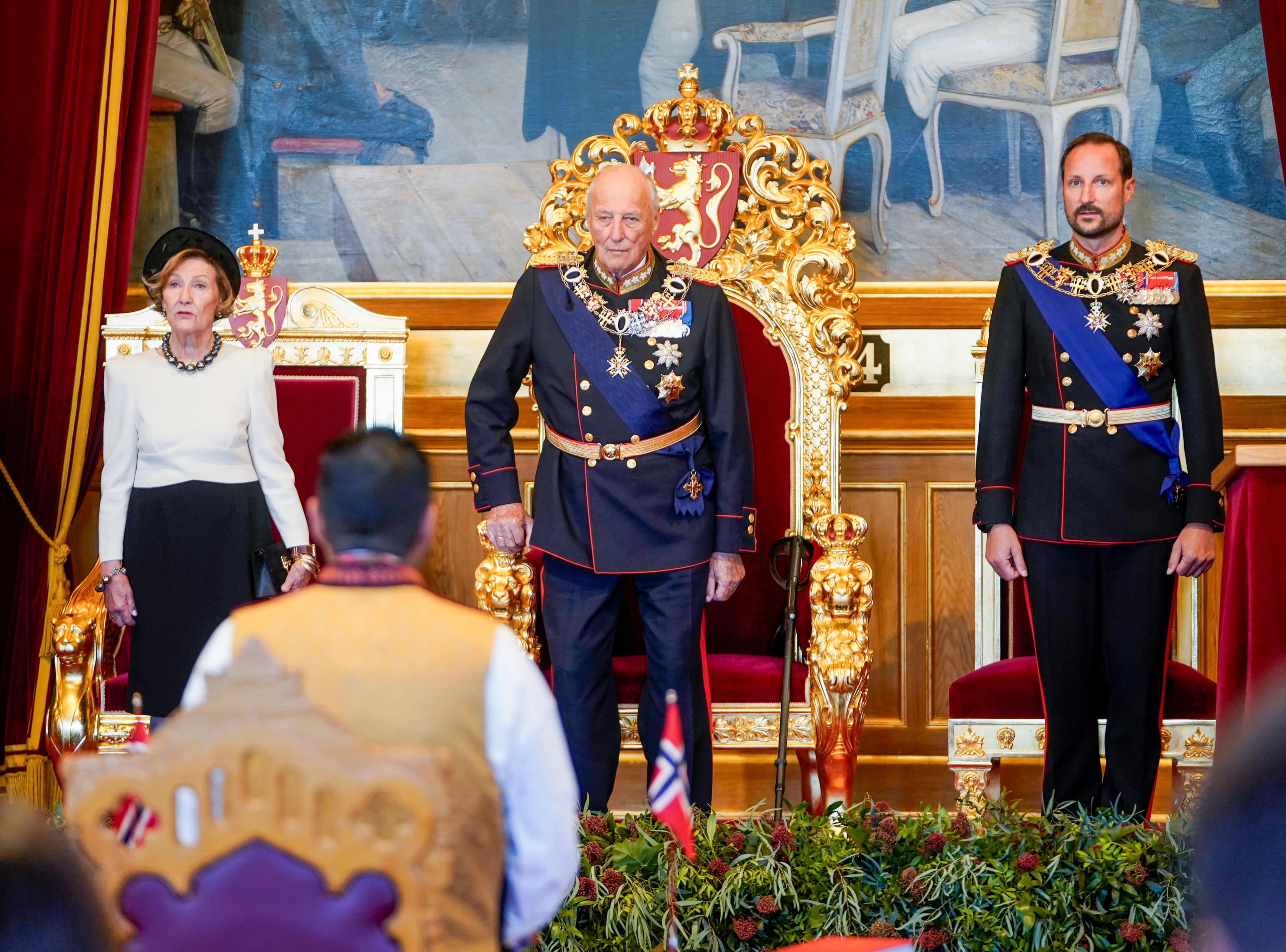 Kong Harald, Dronning Sonja og Kronprins Haakon står ved tronstolen, dronningstolen og prinsestolen i Stortinget.