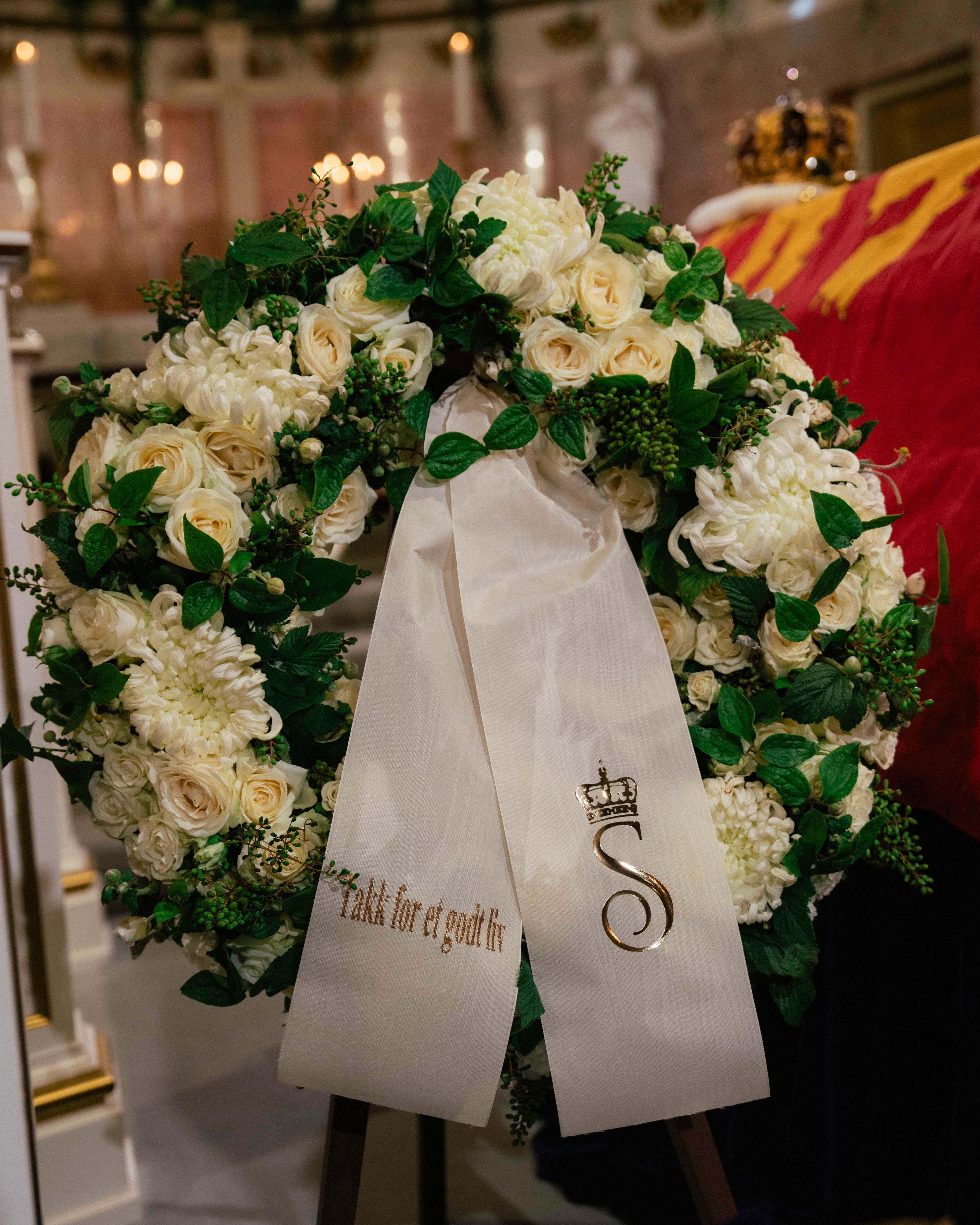 En krans i hvitt og grønt med et hvitt bånd som har påskriften "Takk for et godt liv" og Dronning Sonjas monogram.