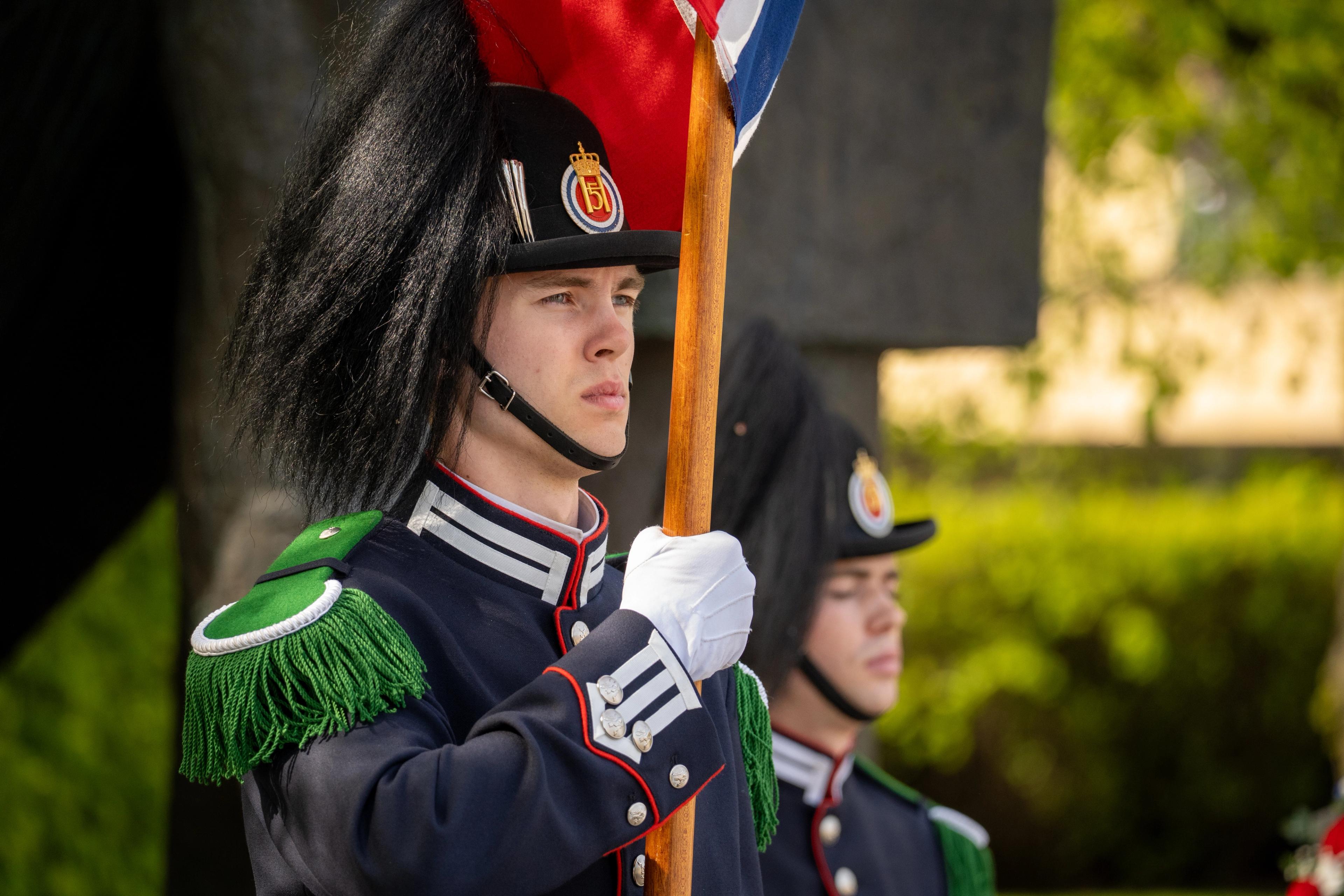 Tett på en gardist som står med stort, norsk flagg ved nasjonalmonumentet.