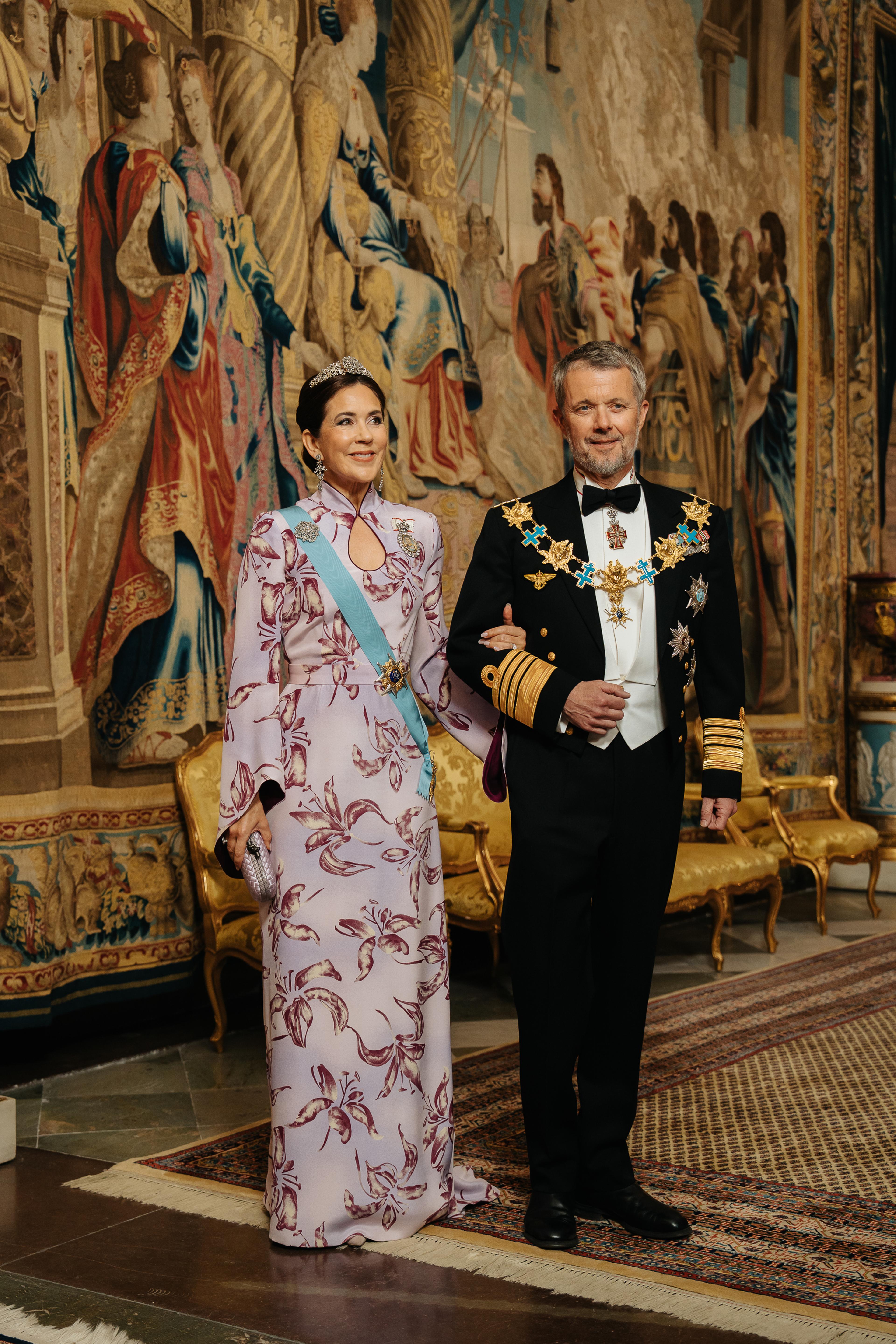 Det danske kongeparet kledd i galla foran et stort veggteppe (gobelin).