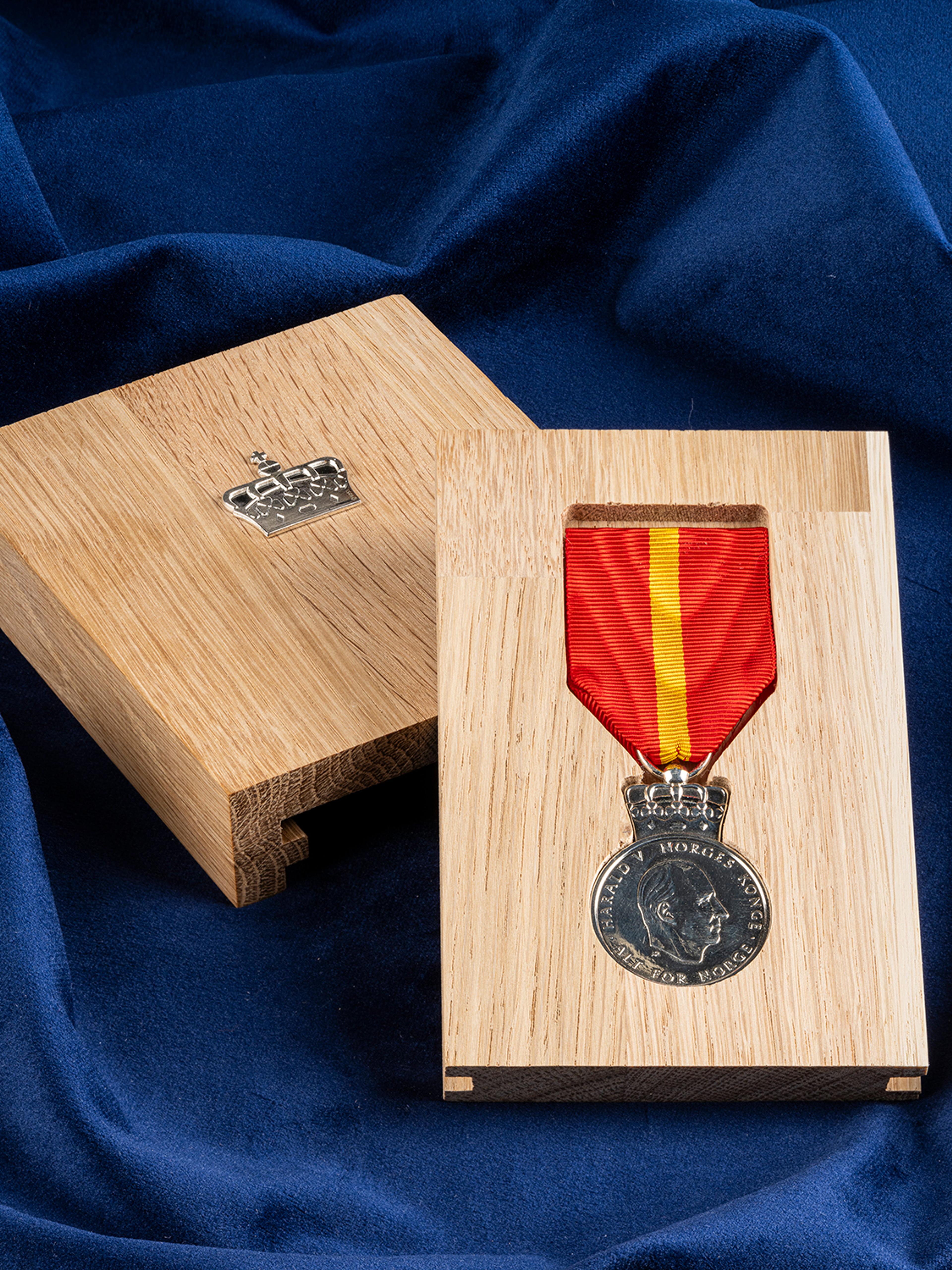 Sølvmedalje med Kongens portrett, fest i et rødt bånd med en gul stripe. Medaljen ligger i et etui av eik plassert på et blått klede.
