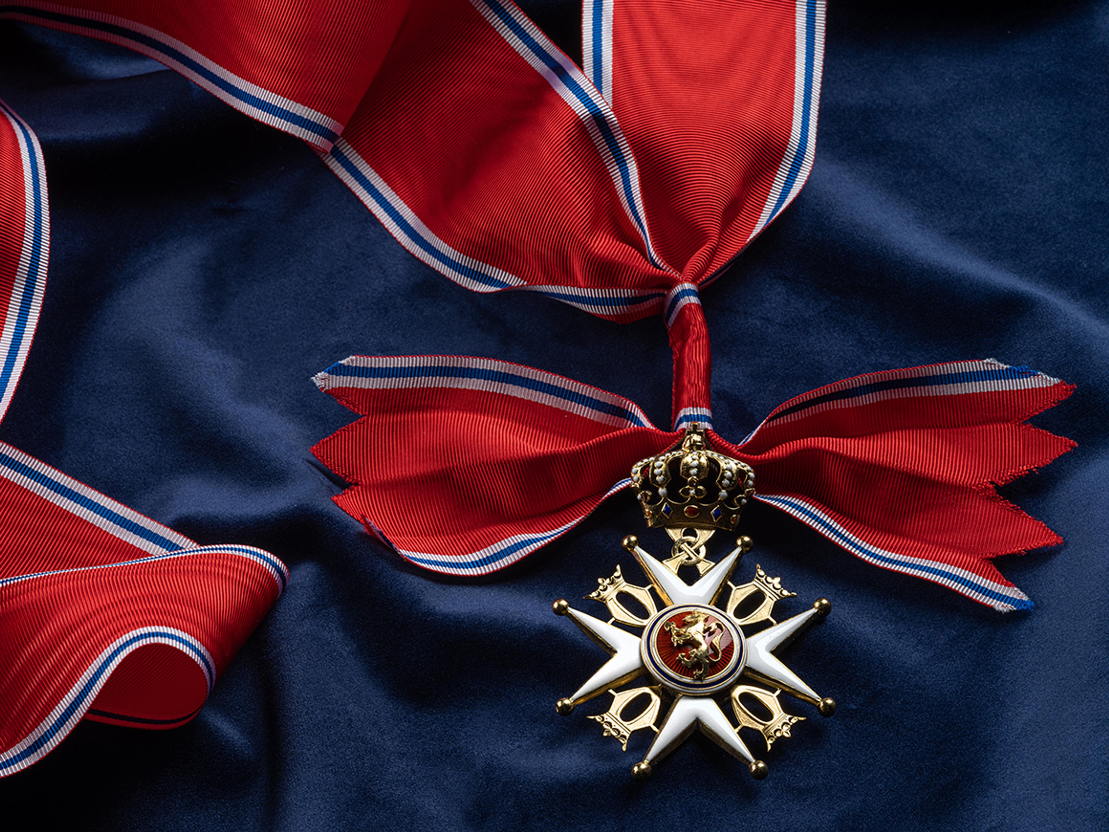 Nærbilde av ordensmerket festet i rødt bånd med blå og hvite striper på en bakgrunn av blått klede.
