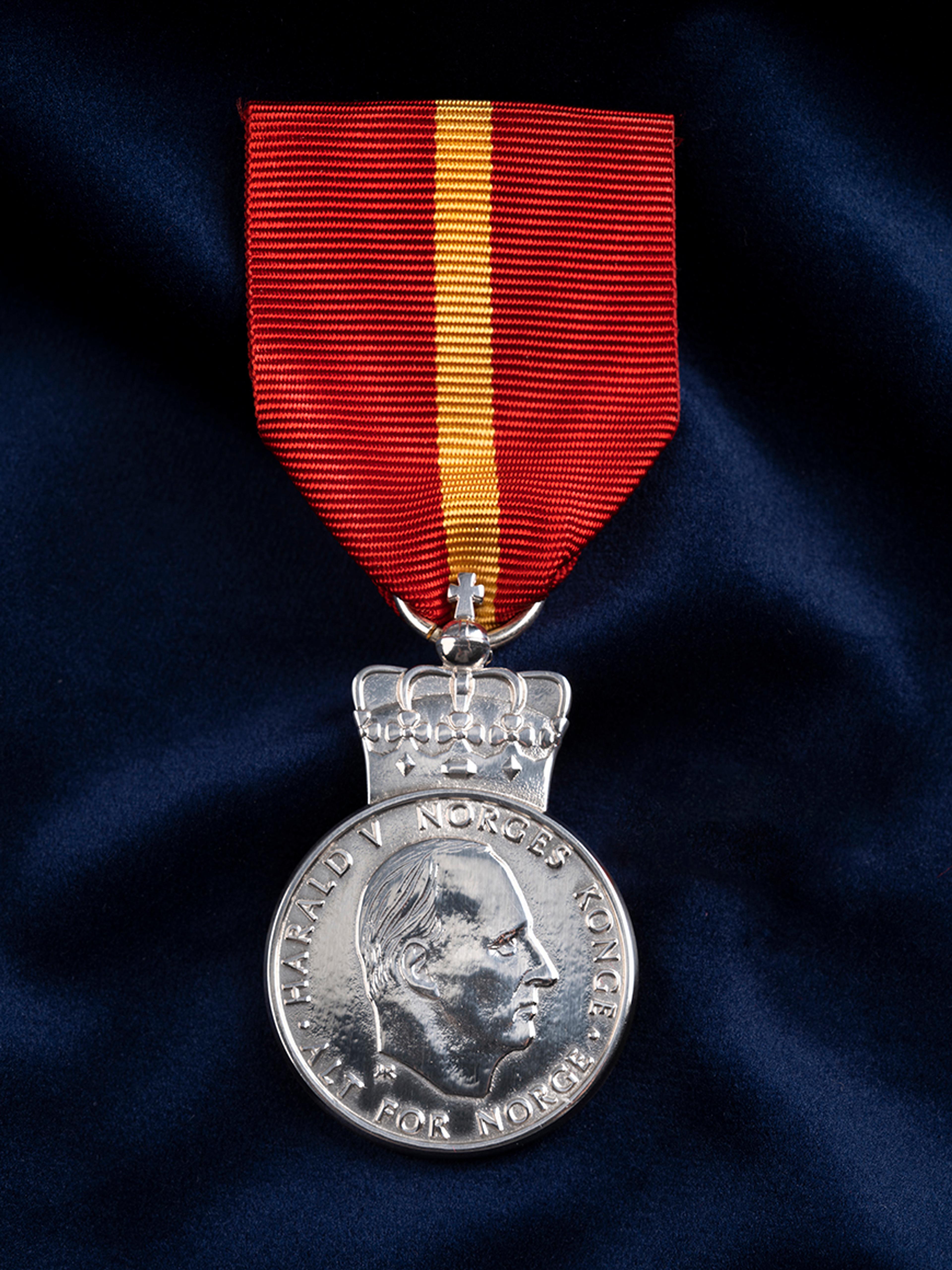 Sølvmedalje med Kongens portrett, festet i et rødt bånd med en gul stripe. Ligger på et dypblått klede.