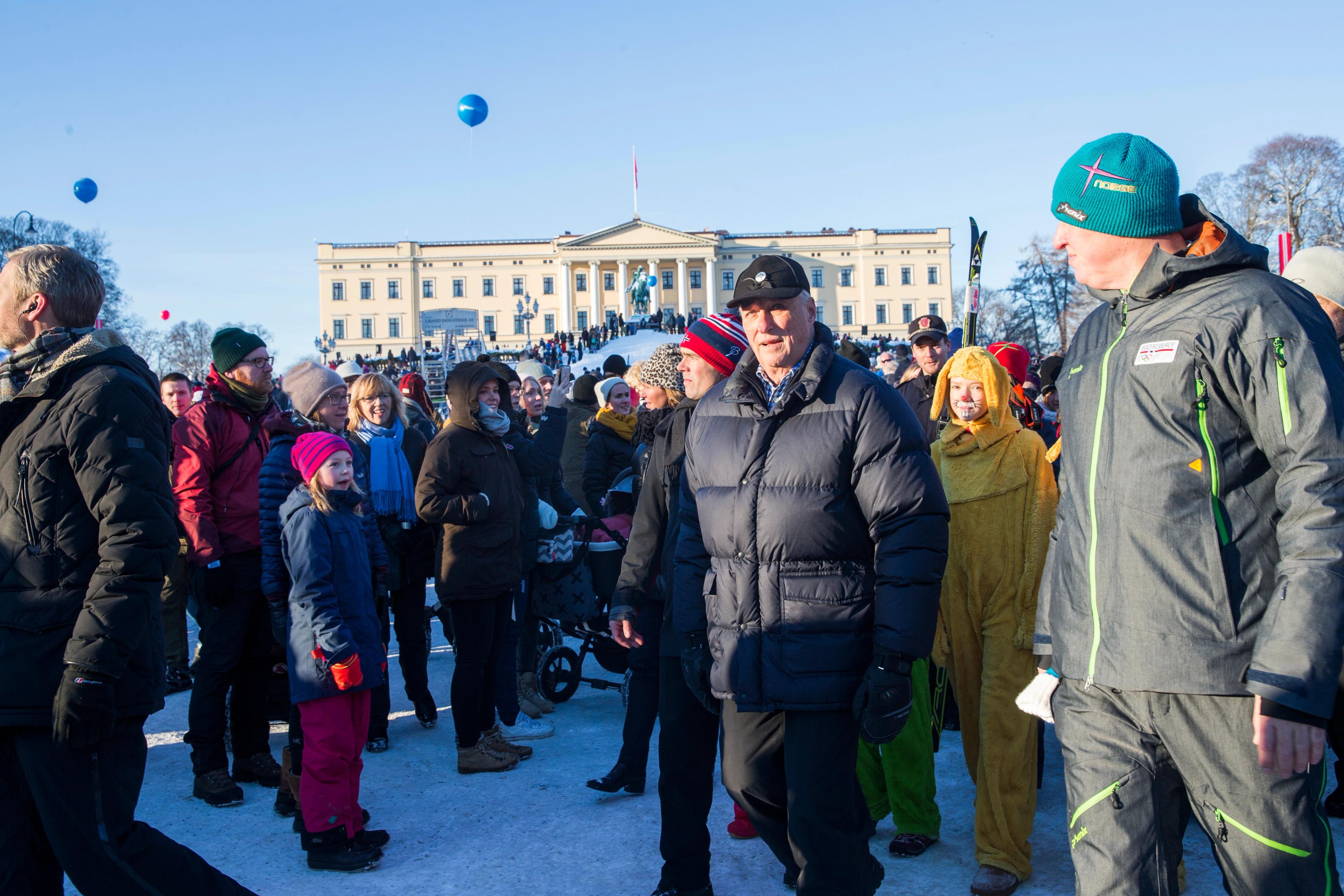 Kong Harald i vintertøy ute blant folk på Slottsplassen. Bak ham ses Slottet med akebakke foran og blå ballonger.