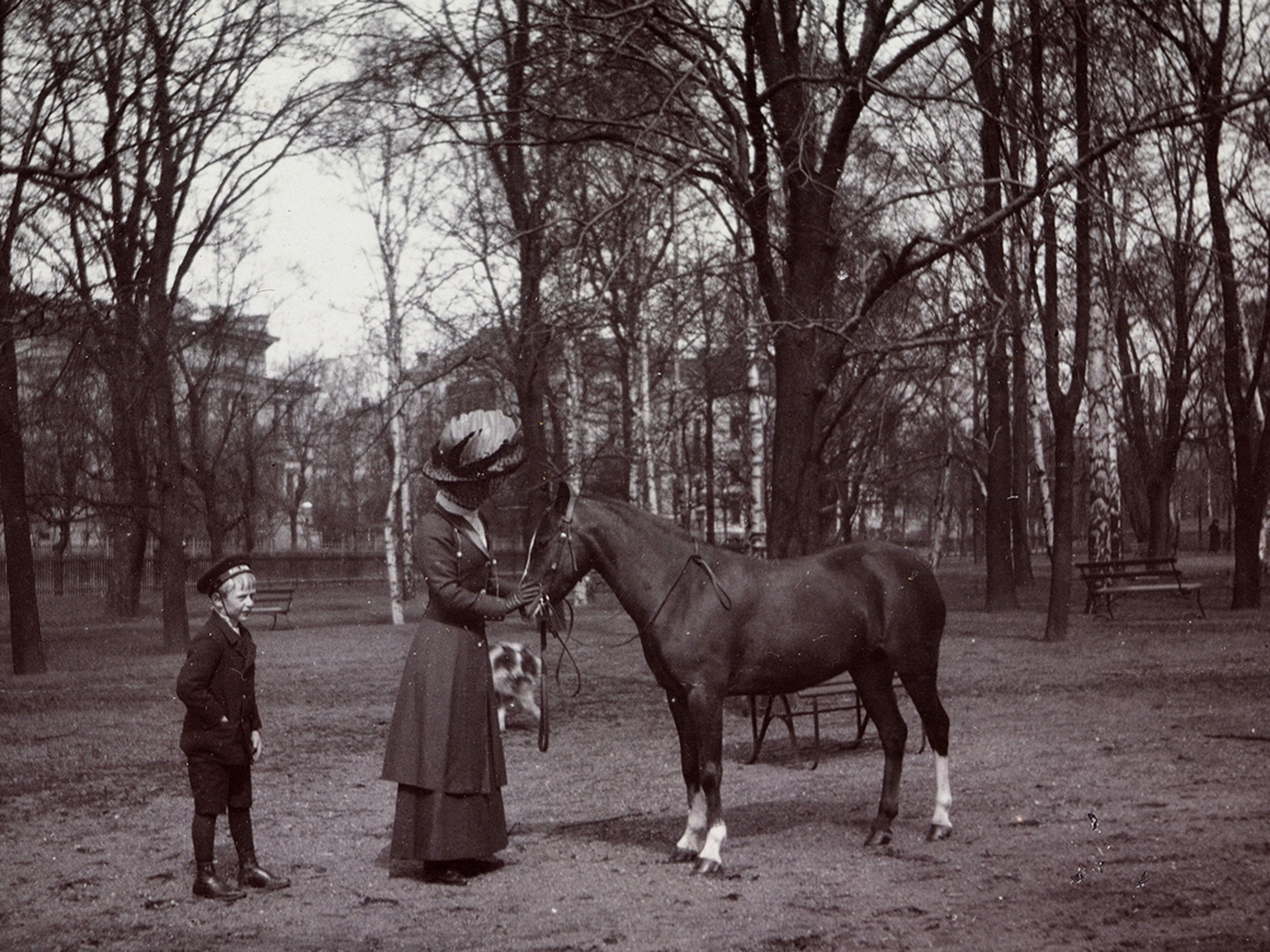 Dronning Maud klapper hesten på mulen. Kronprins Olav står litt bak henne.