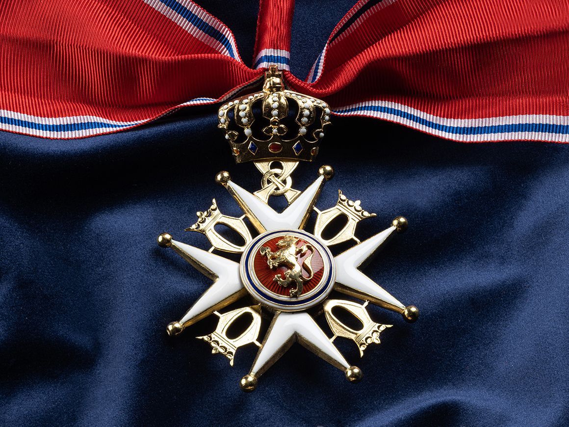 Nærbilde av ordensmerket festet i rødt bånd med blå og hvite striper på en bakgrunn av blått klede.