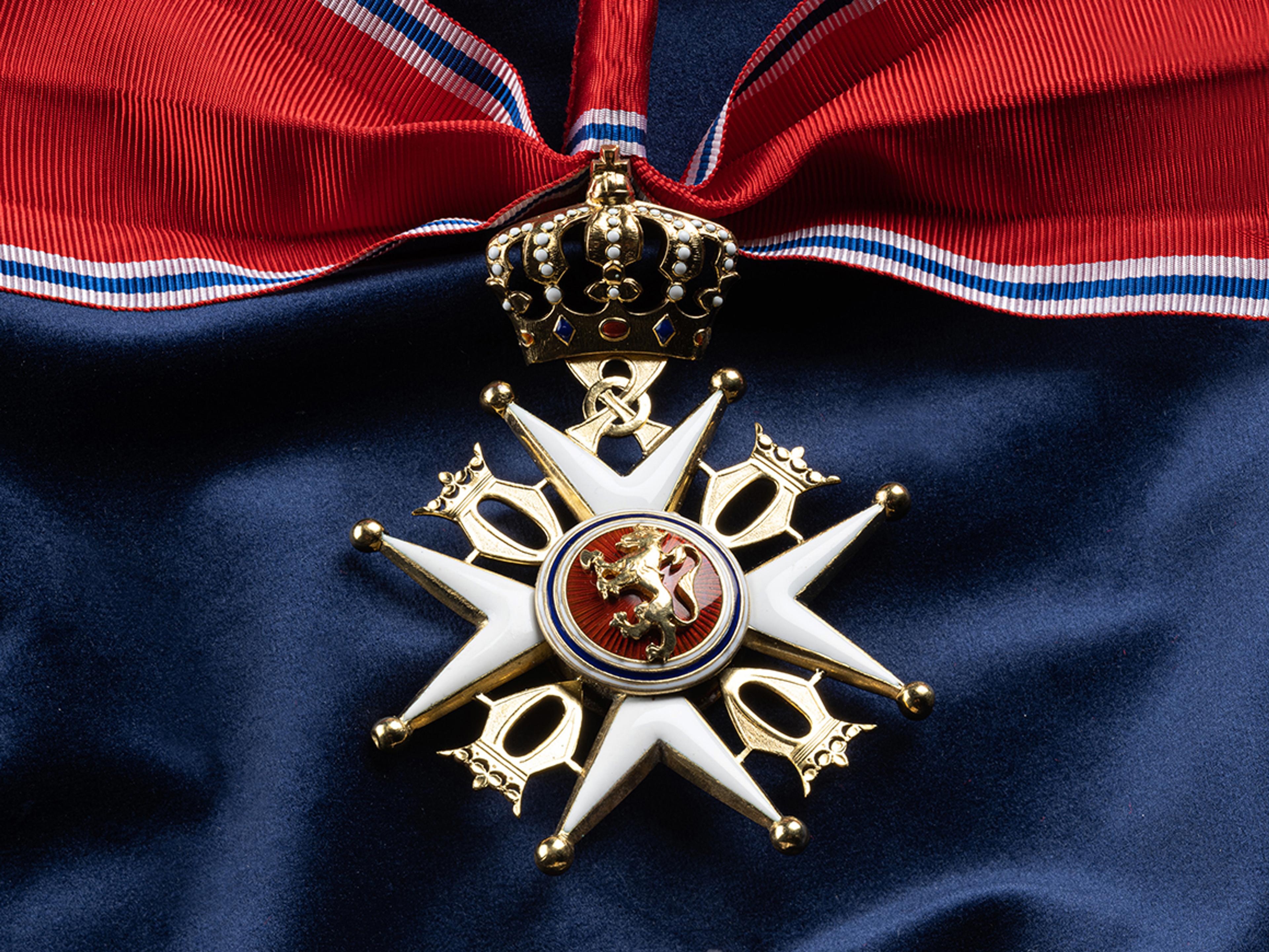 Nærbilde av ordensmerket festet i rødt bånd med blå og hvite striper på en bakgrunn av blått klede.