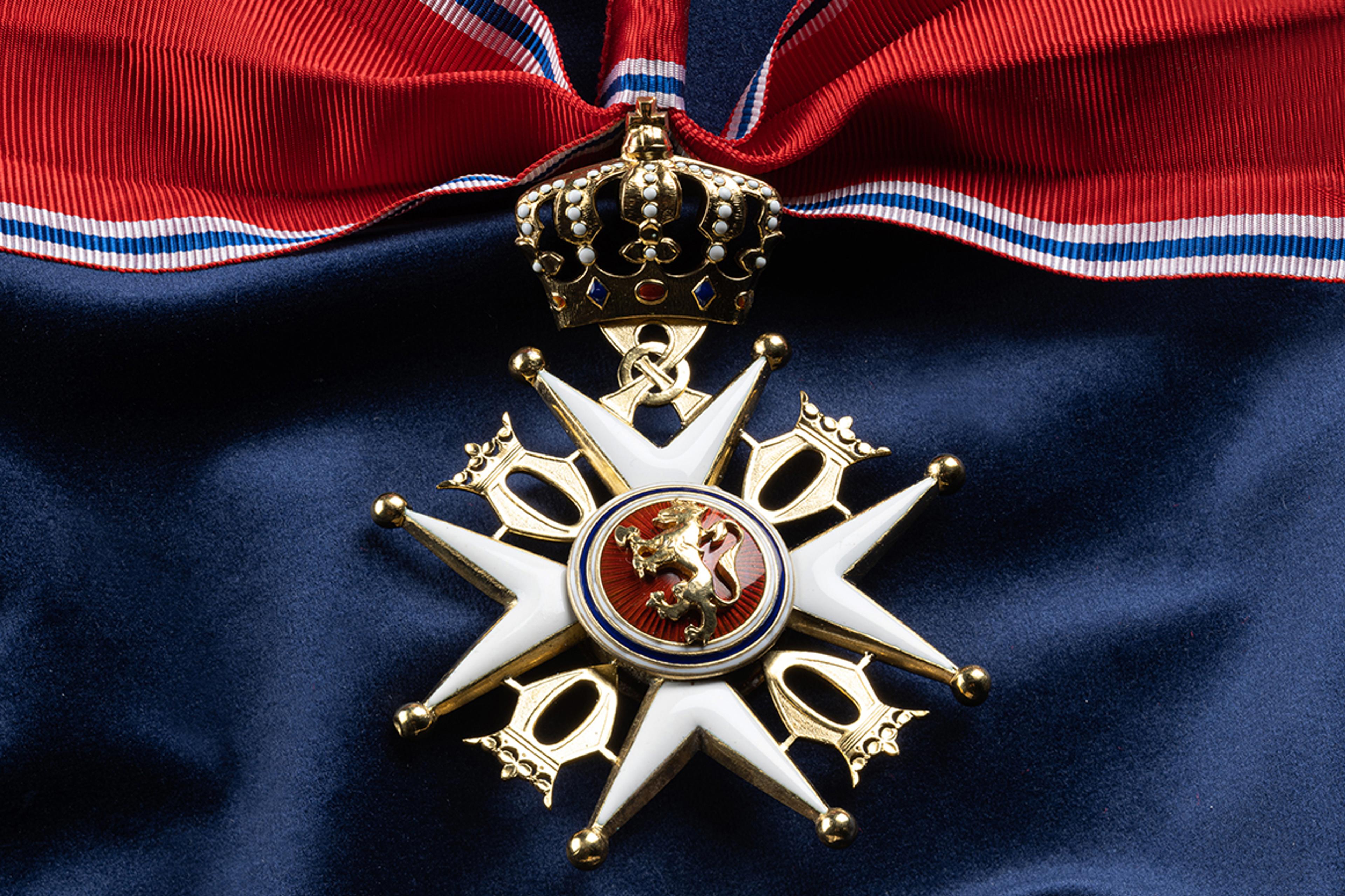Nærbilde av ordensmerket festet i rødt bånd med blå og hvite striper på en bakgrunn av blått klede.
