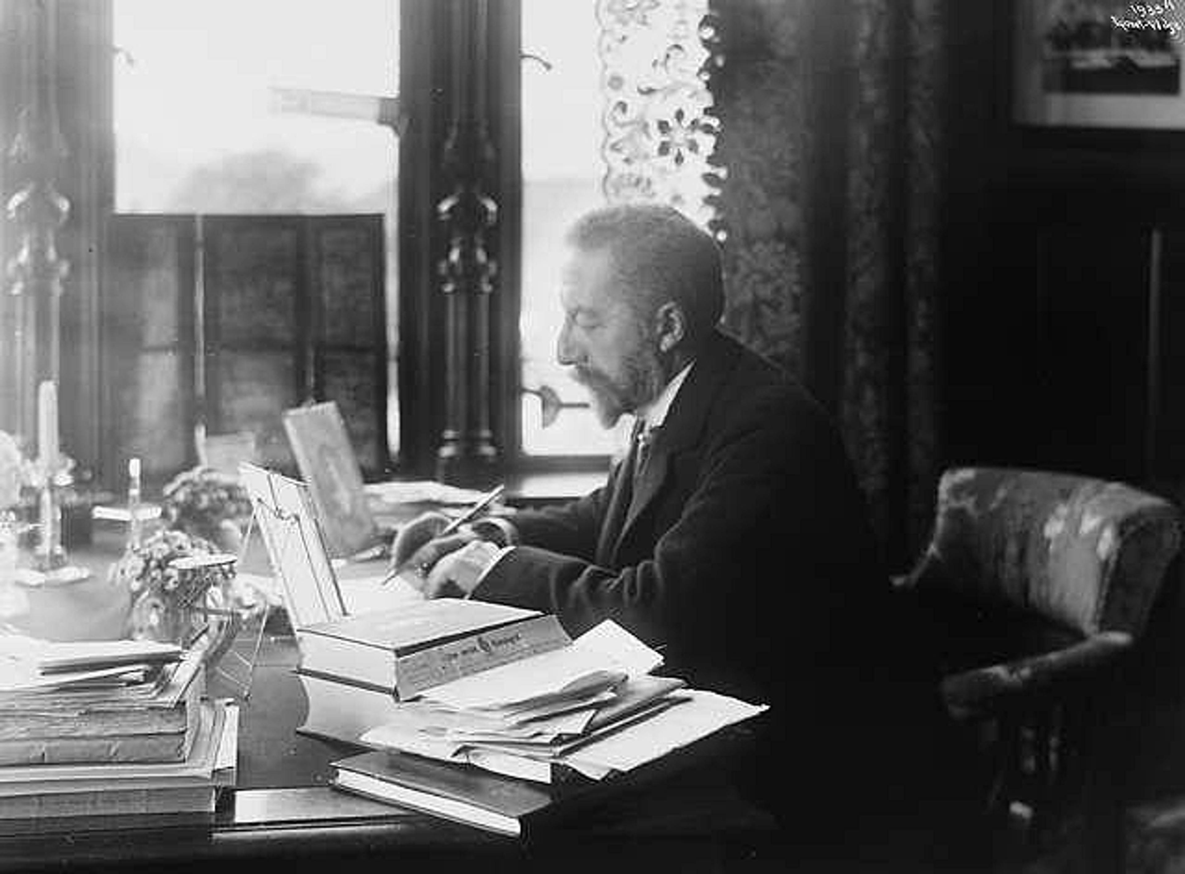 Michelsen sitter og skriver ved et skrivebord fullt av bøker og papirer.