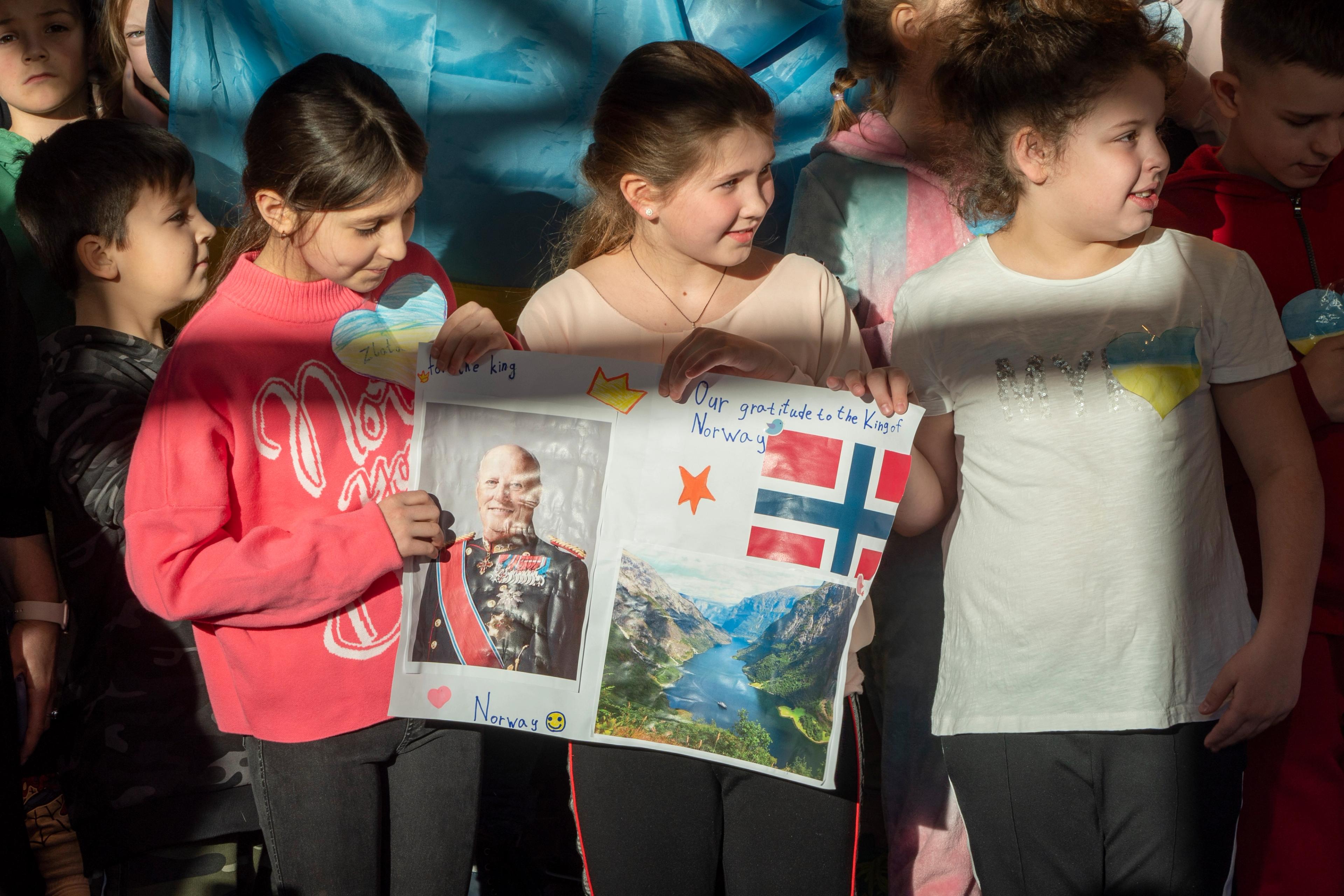 Tre jenter holder i en plakat med bilde av Kong Harald og teksten "Our gratitude to the King of Norway". De har papirhjerter festet på genserne i fargene til det ukrainske flagget - gult og blått.