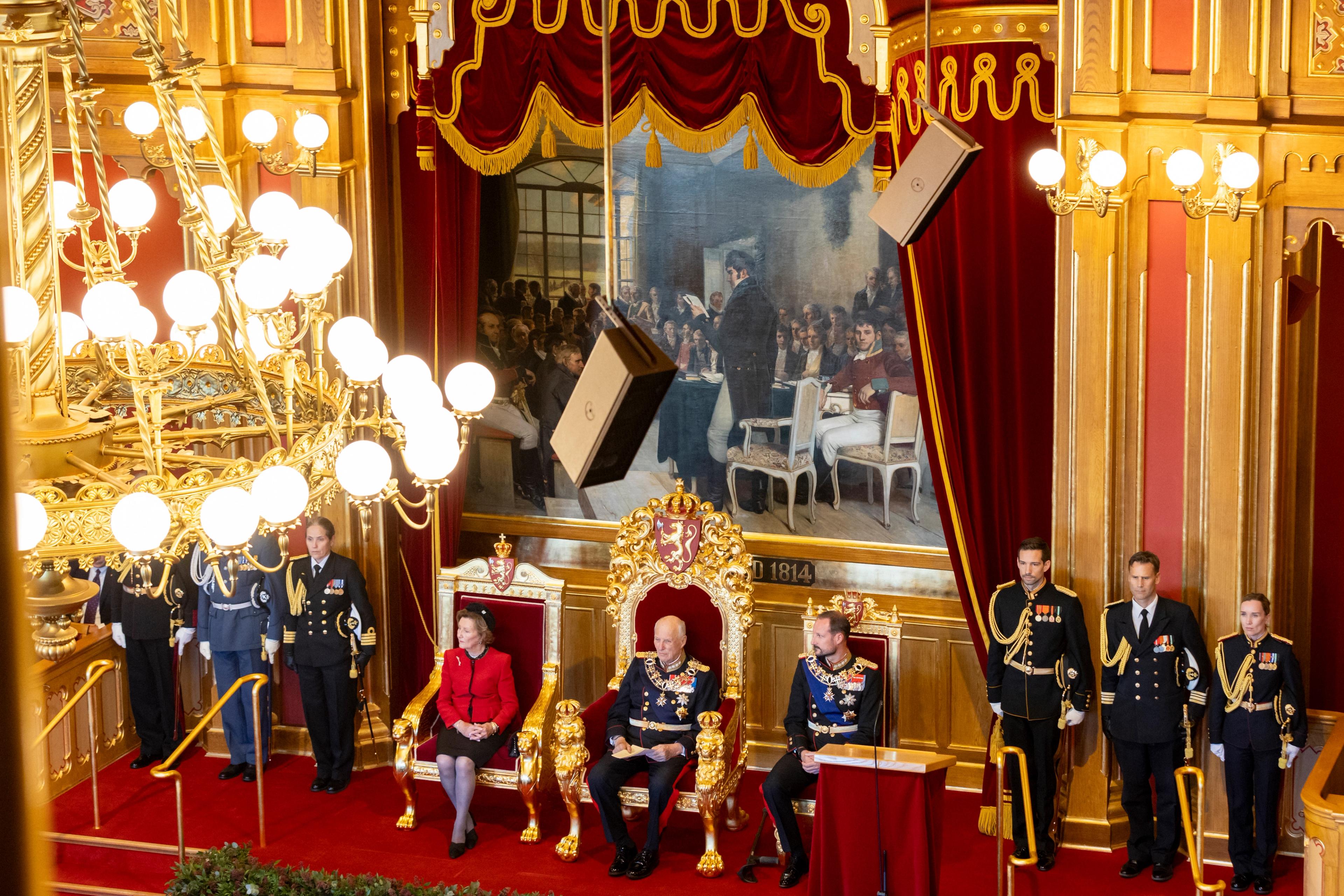 Kong Harald, Dronning Sonja og Kronprins Haakon sitter i stortingssalen med tre adjutanter stående på høyre side og tre overadjutanter stående på venstre side.