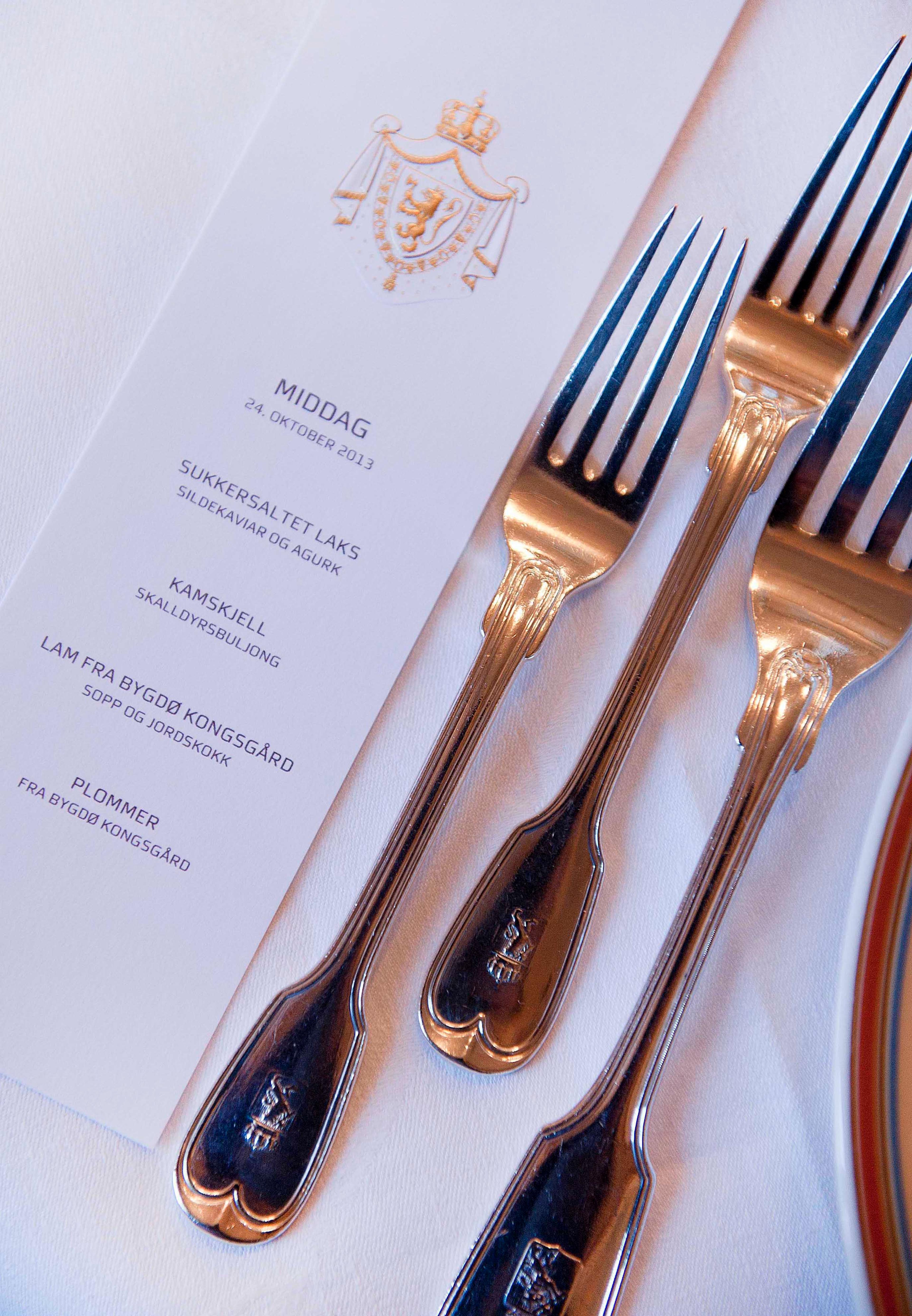 En smal meny på, tykt hvitt papir og med Kongevåpenet i gull øverst, beskriver kveldens meny - laks, kamskjell, lam og plommer.
