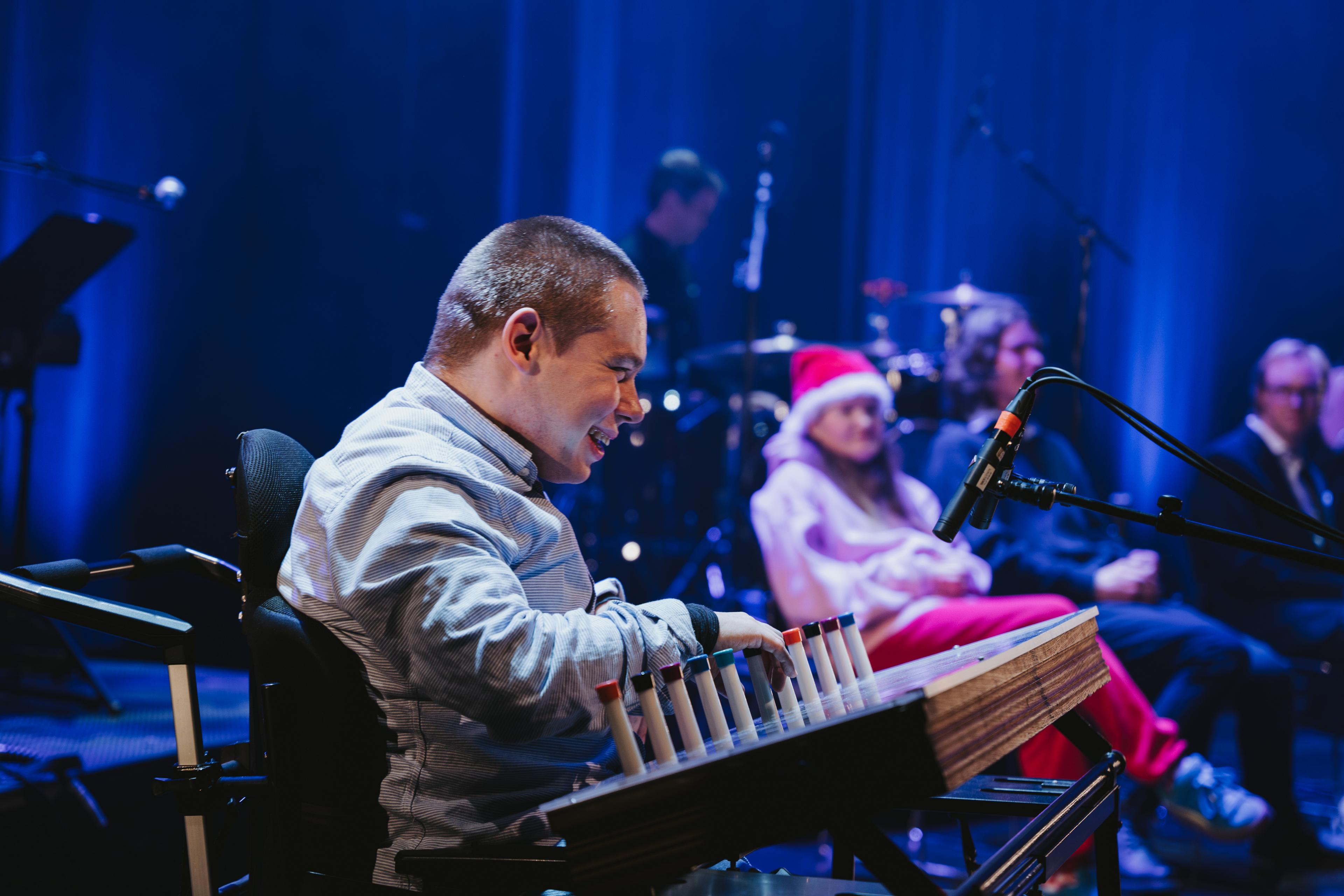 Med eit breitt smil spelar ein mann i rullestol på eit klanginstrument på ei scene.